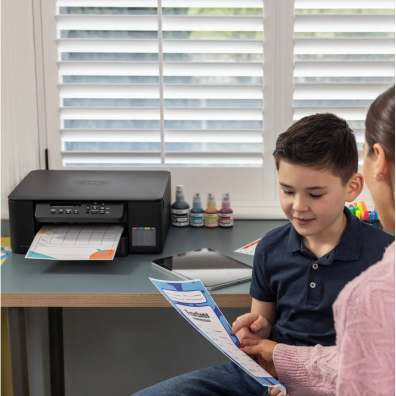 A boy and woman look at a printed paper. A black printer sits on a desk next to ink bottles. Window in the background.