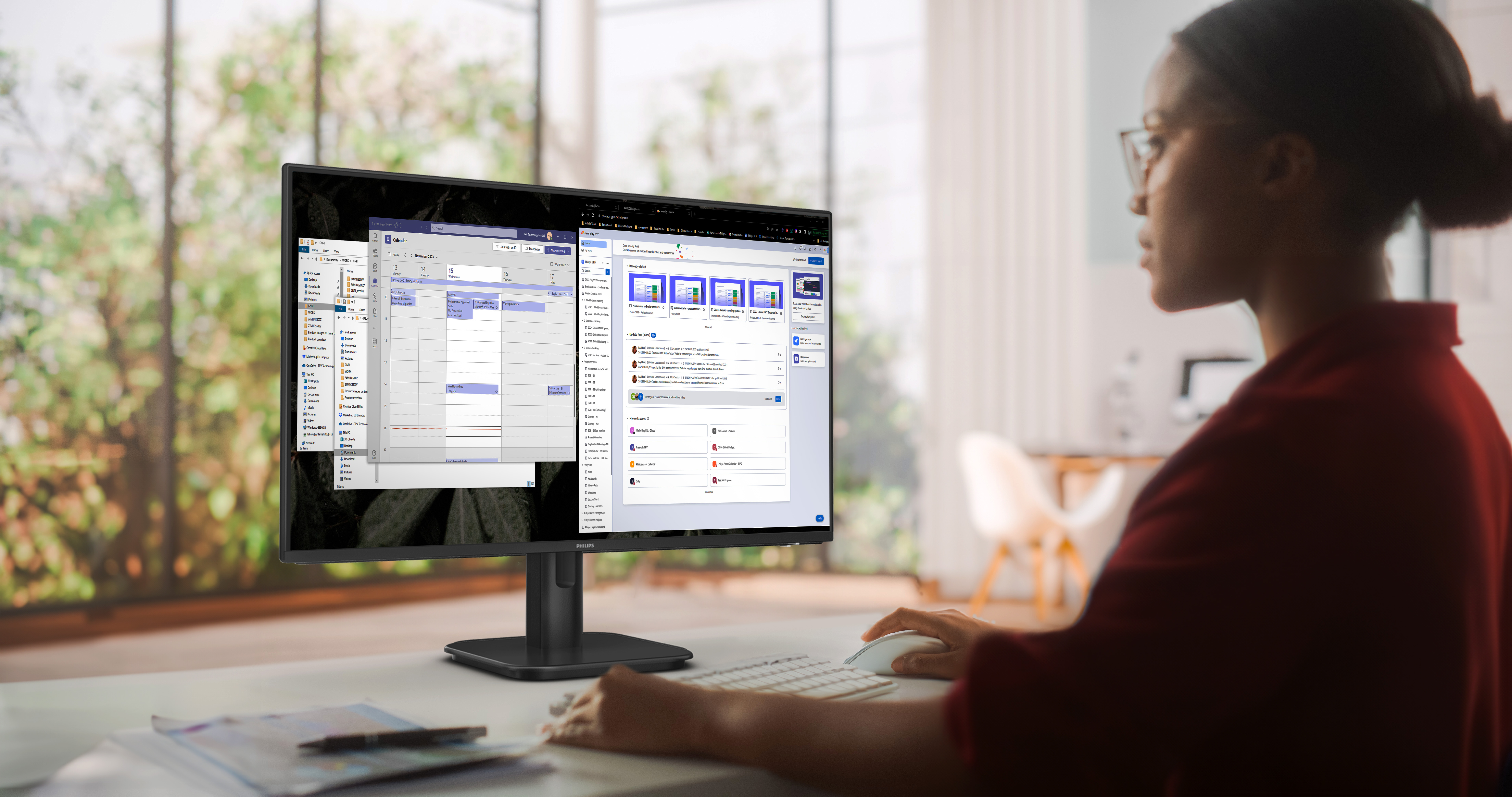 Frau benutzt Computer mit Kalender auf dem Bildschirm. Sie trägt Brille und ein rotes Hemd, drinnen, weißer Schreibtisch.