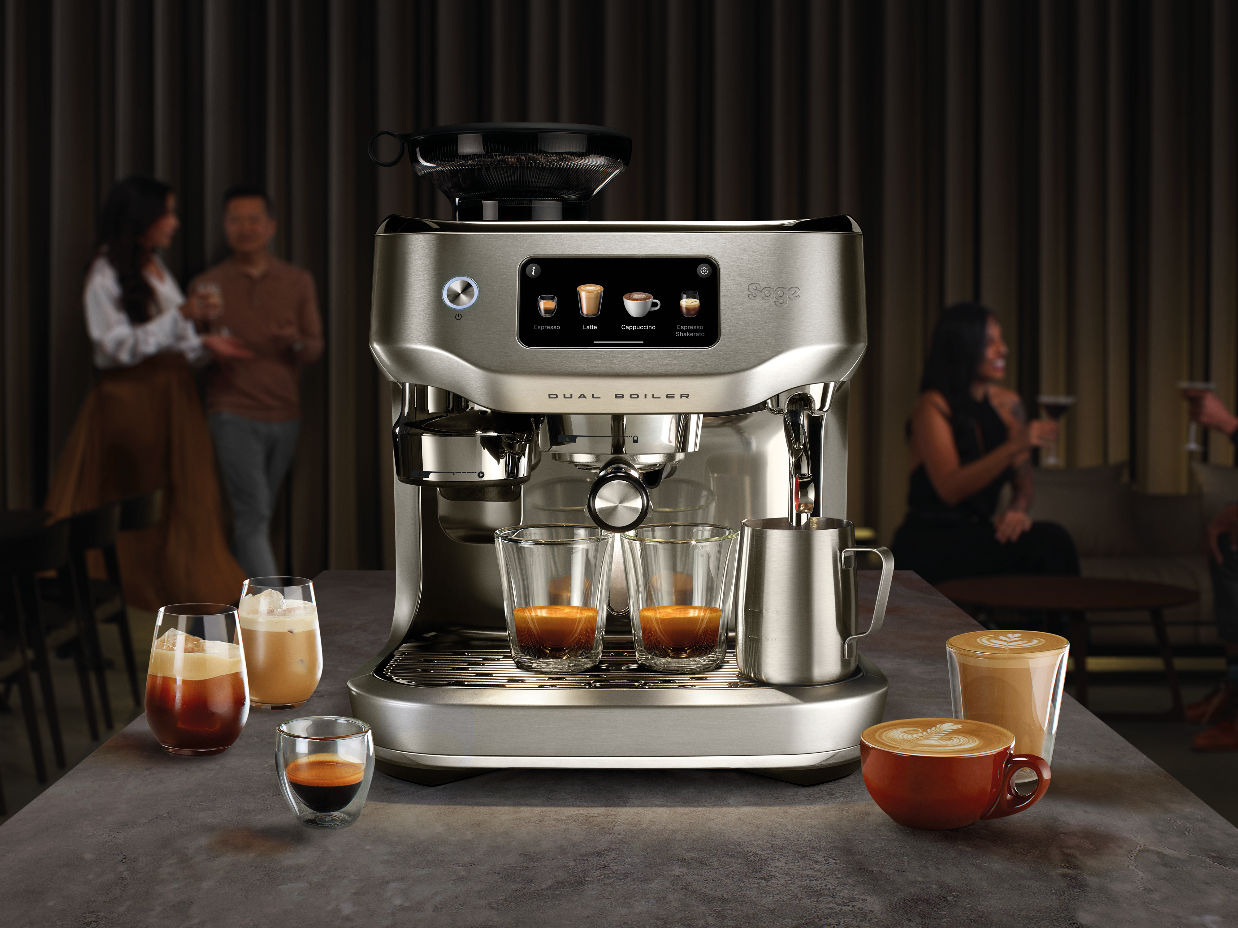 Eine silberne Espressomaschine mit Kaffeegetränken und Personen im Hintergrund. Auf dem Tisch stehen verschiedene Kaffeebecher und Gläser.
