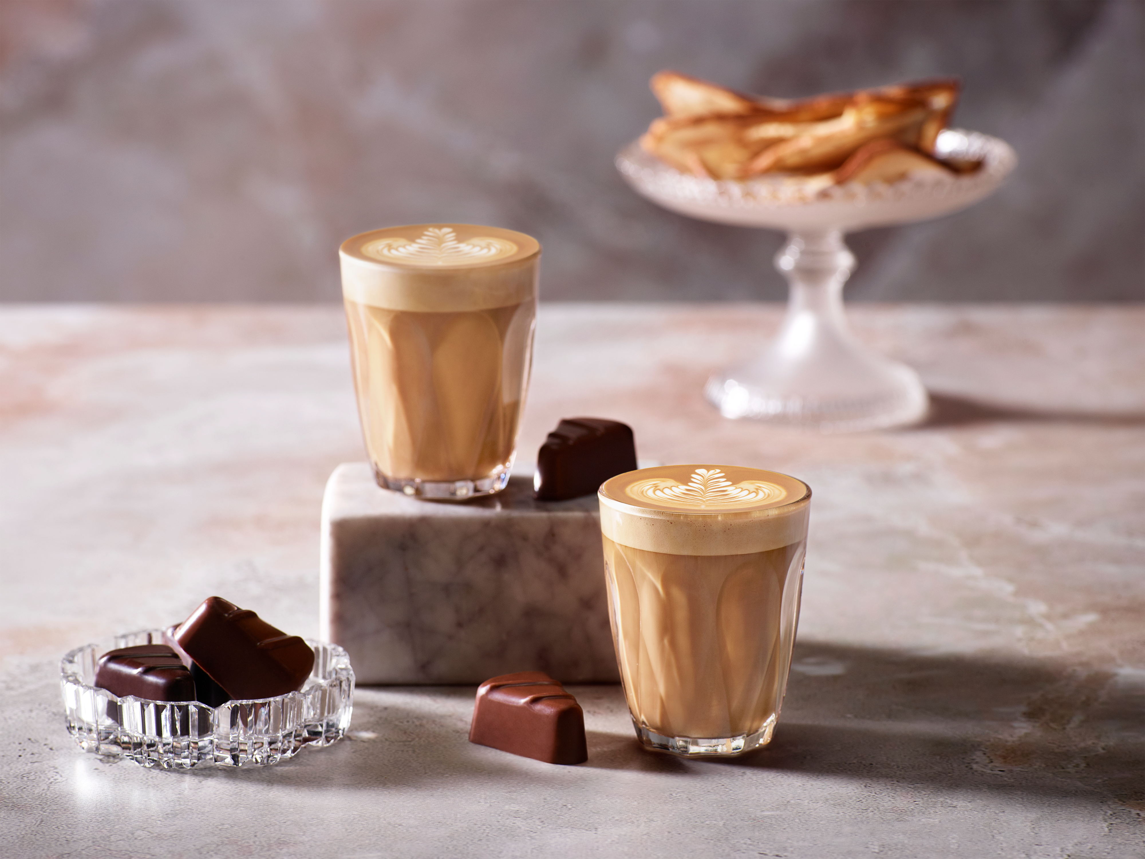 Zwei Latte Macchiatos mit Latte Art, Pralinen und ein Teller Gebäck auf einer Marmoroberfläche.