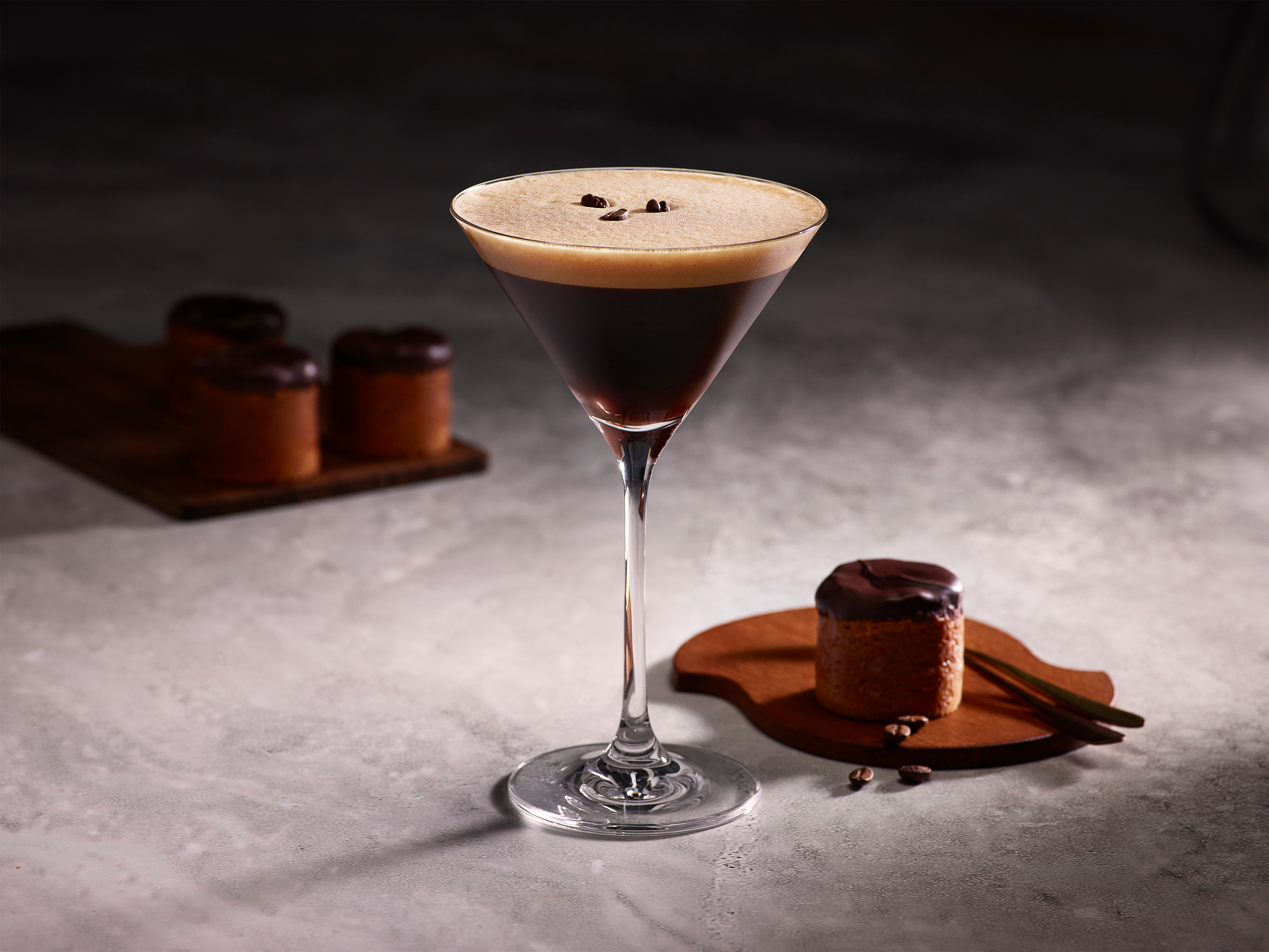 Ein Martiniglas mit Espresso, garniert mit Schaum und Kaffeebohnen. Kuchenstücke auf Holzbrettern befinden sich im Hintergrund.