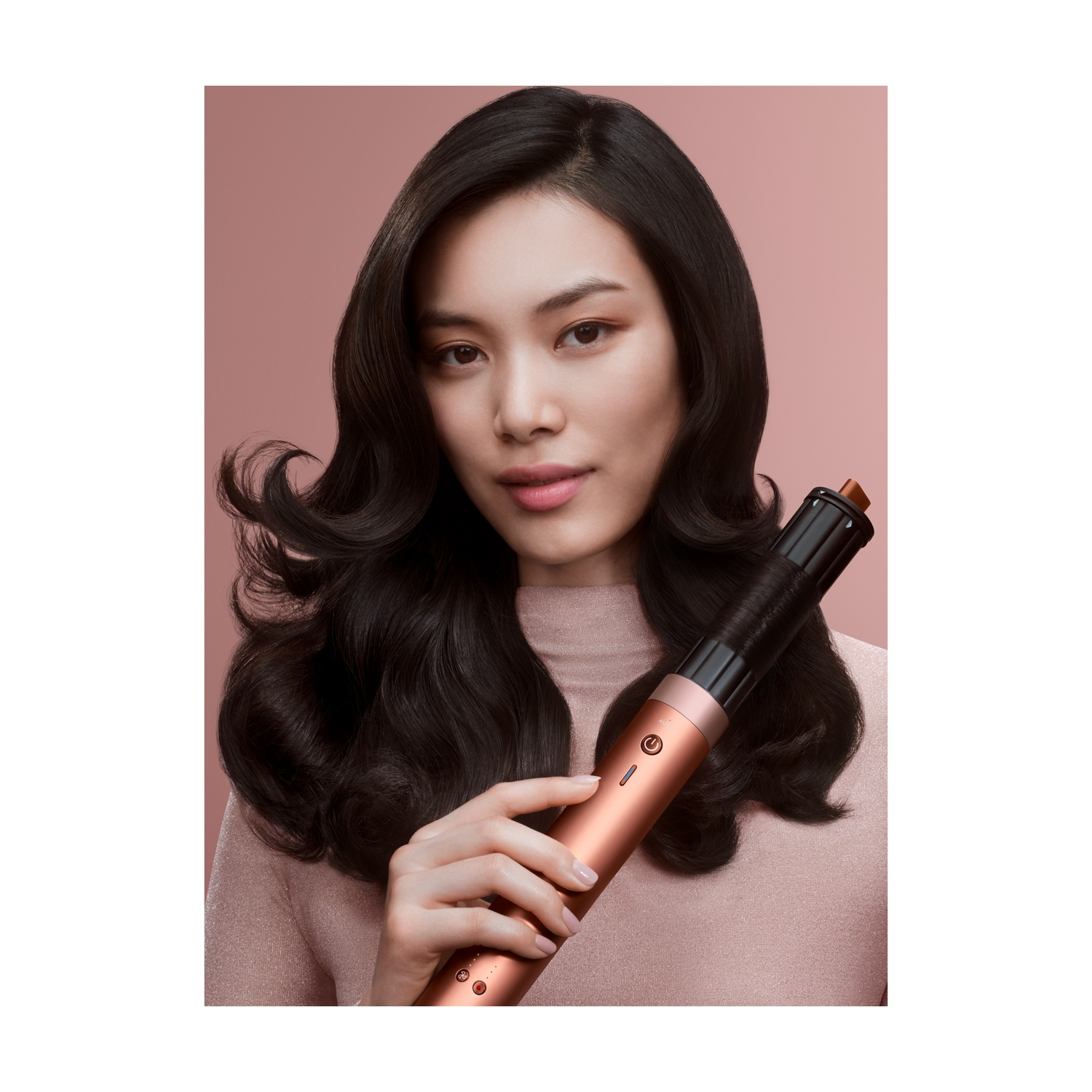 Una donna con i capelli scuri tiene in mano uno strumento per lo styling dei capelli. Indossa un top rosa, con sfondo rosa. Lo strumento è color rame e nero.