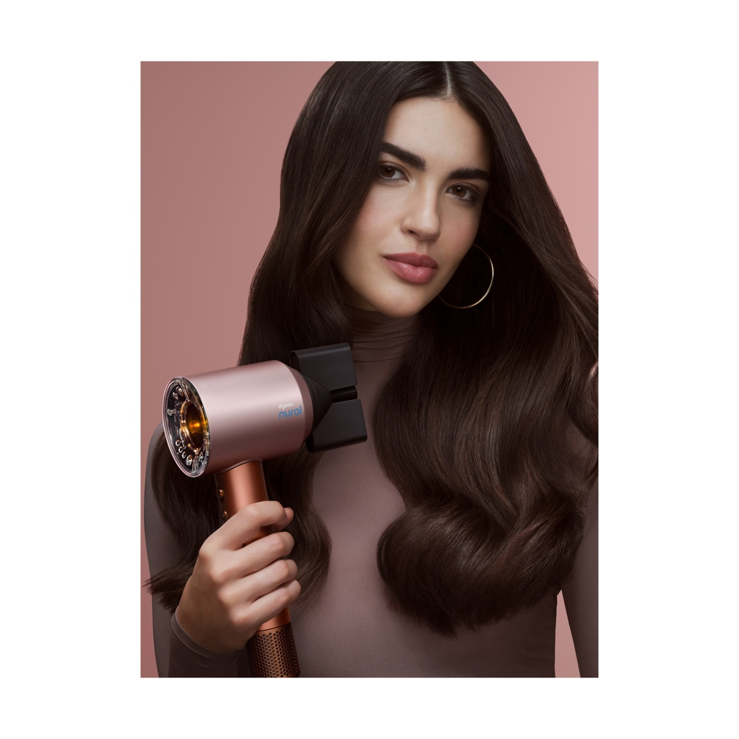 Una donna tiene in mano un asciugacapelli color oro rosa. Ha lunghi capelli scuri e indossa un top marrone. Lo sfondo è rosa chiaro.