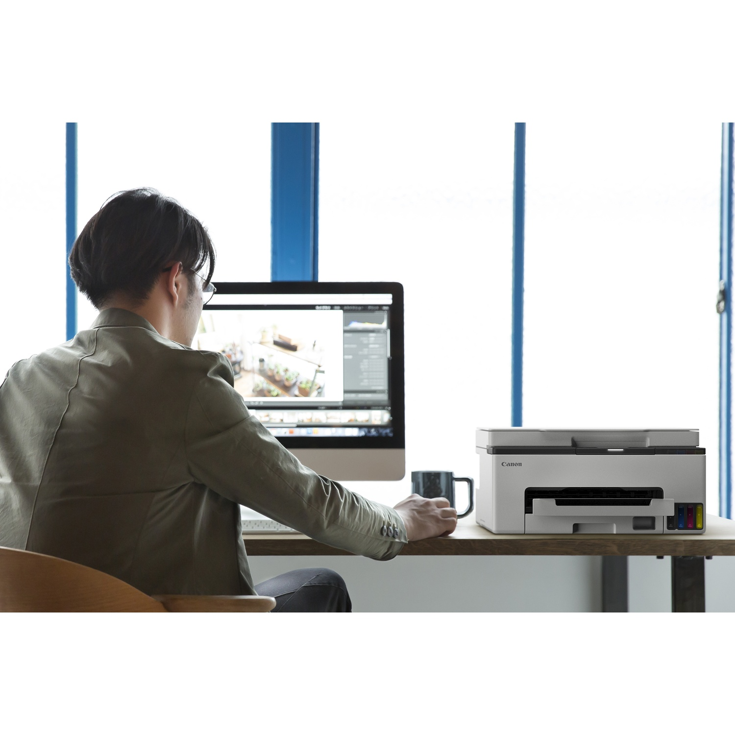 Un homme est assis à un bureau, utilisant un ordinateur. Une imprimante Canon est sur le bureau. Une tasse est à proximité. L'arrière-plan montre une fenêtre.