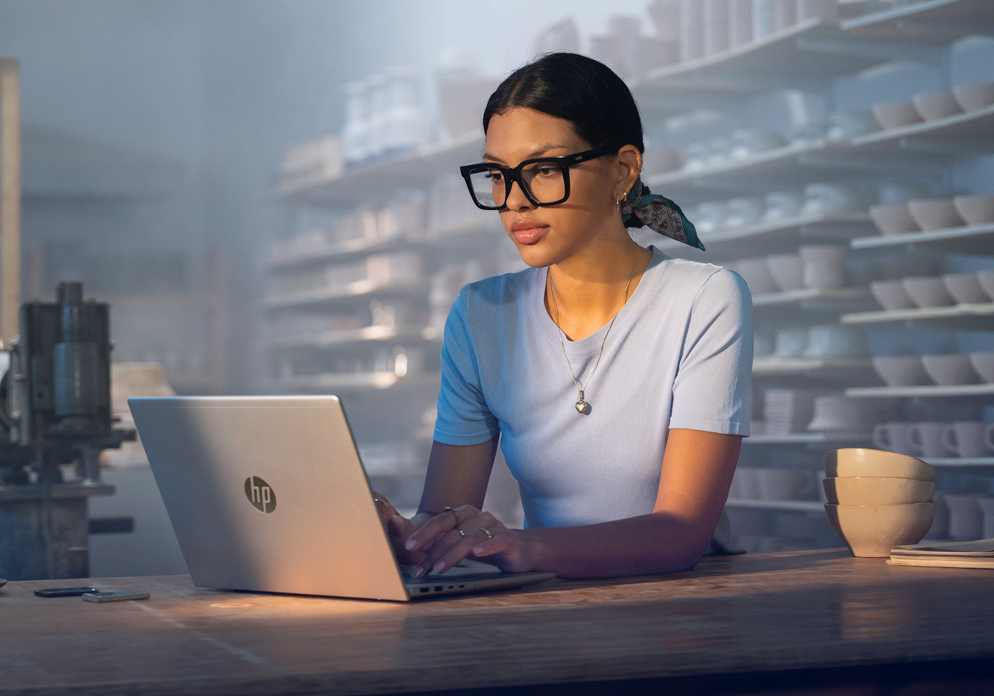 Een vrouw met een bril gebruikt een HP-laptop in een pottenbakkersstudio. Schappen met aardewerk op de achtergrond.