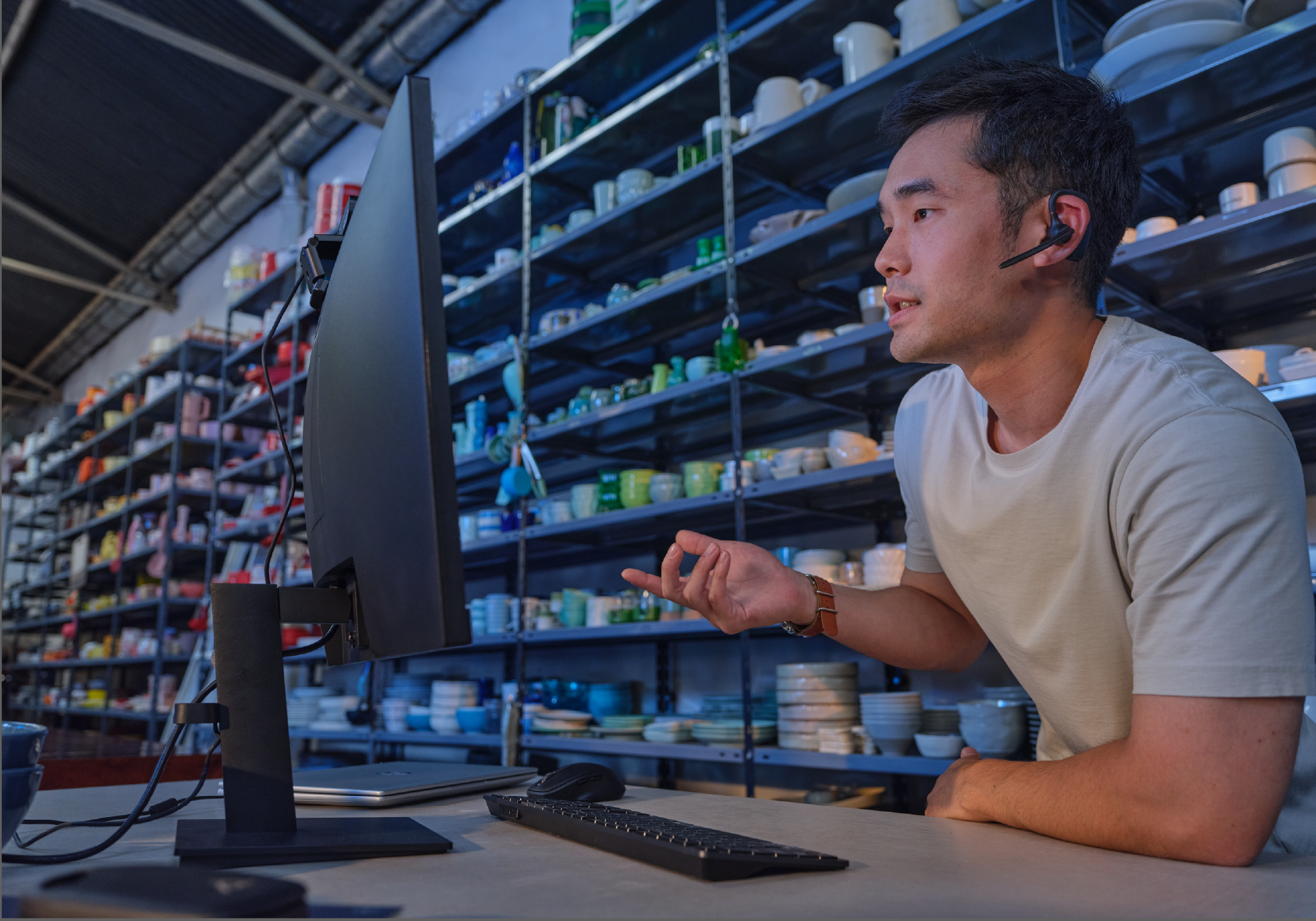 A man with a headset gestures at a computer screen. Shelves filled with pottery are in the background. A keyboard and laptop are on the desk.