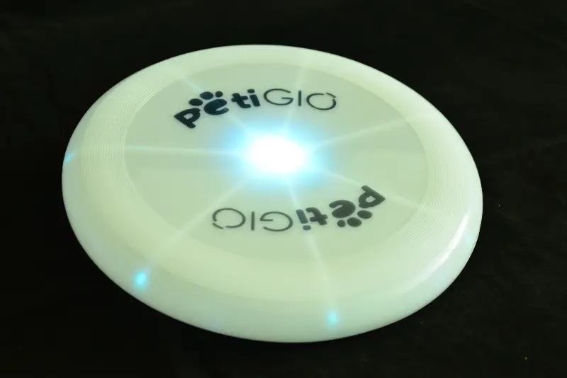Biały frisbee z tekstem 'pëtiGIO' i niebieskimi światłami, wyśrodkowany na czarnej powierzchni. Frisbee jasno świeci w środku.