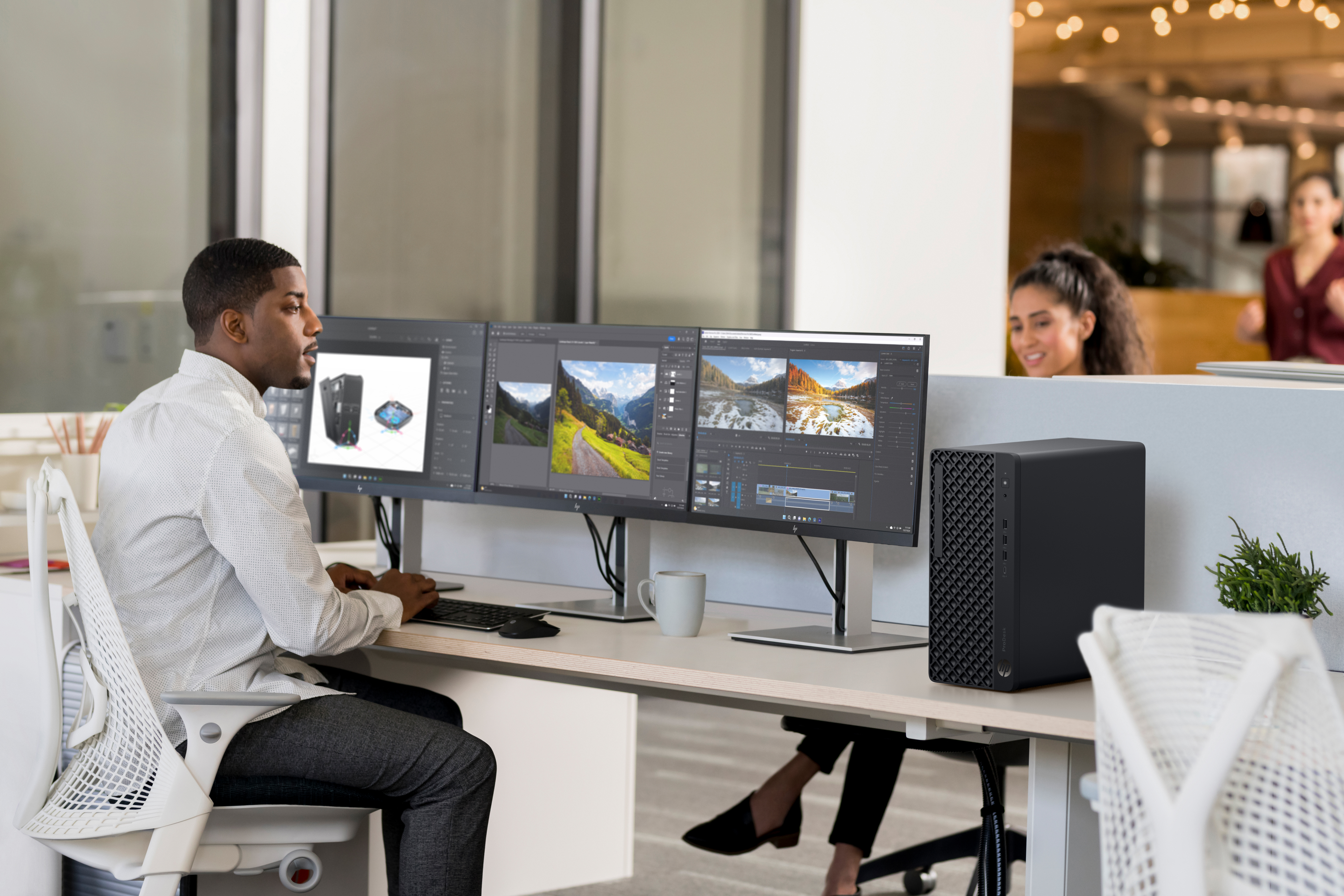 Ein Mann arbeitet an einem Schreibtisch mit drei Monitoren, auf denen Bilder angezeigt werden. Ein schwarzer Computer steht daneben. Andere Leute sind im Hintergrund.