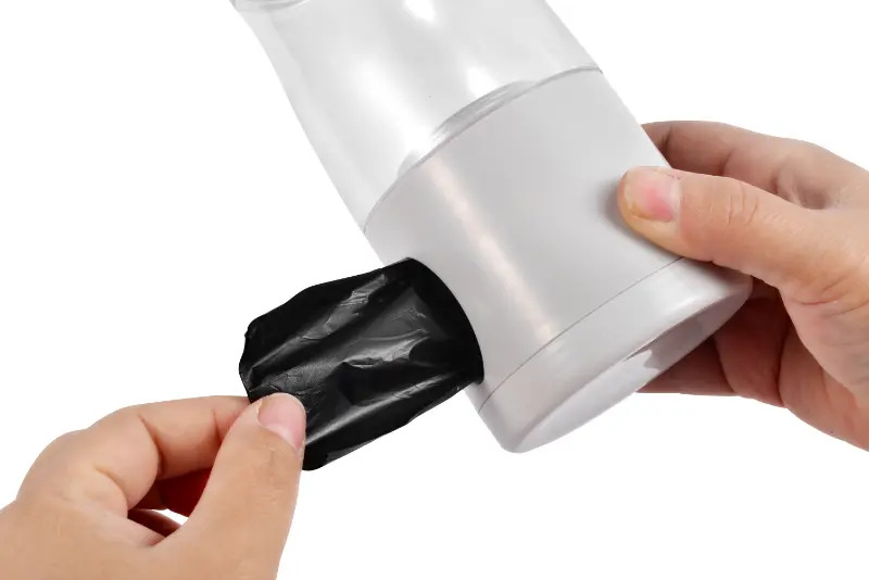 Ręce trzymają biały i przezroczysty dozownik z czarną wyciągniętą torbą. Tło jest białe.