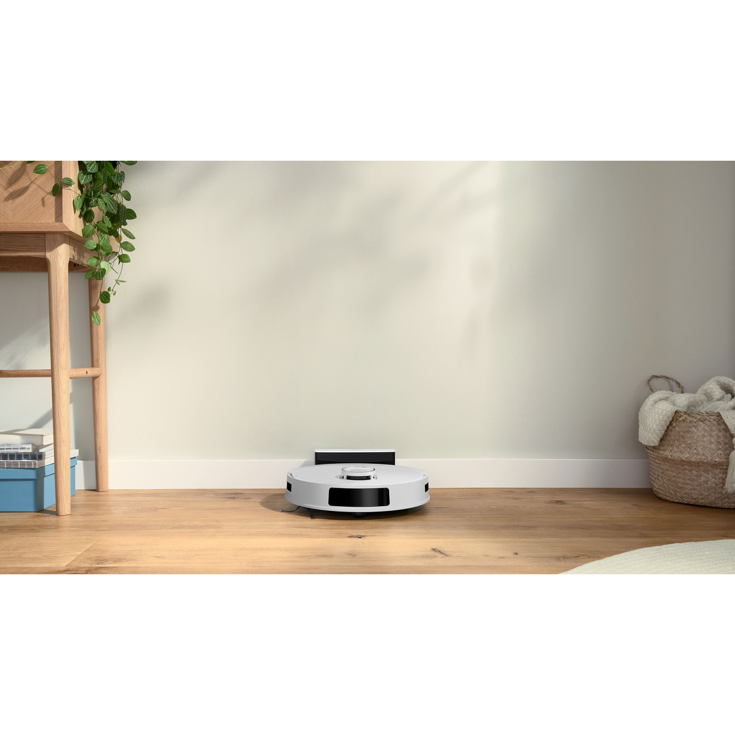 Un aspirapolvere robotico bianco su un pavimento in legno. Si vedono un cesto con una coperta, una mensola in legno e una pianta pensile.