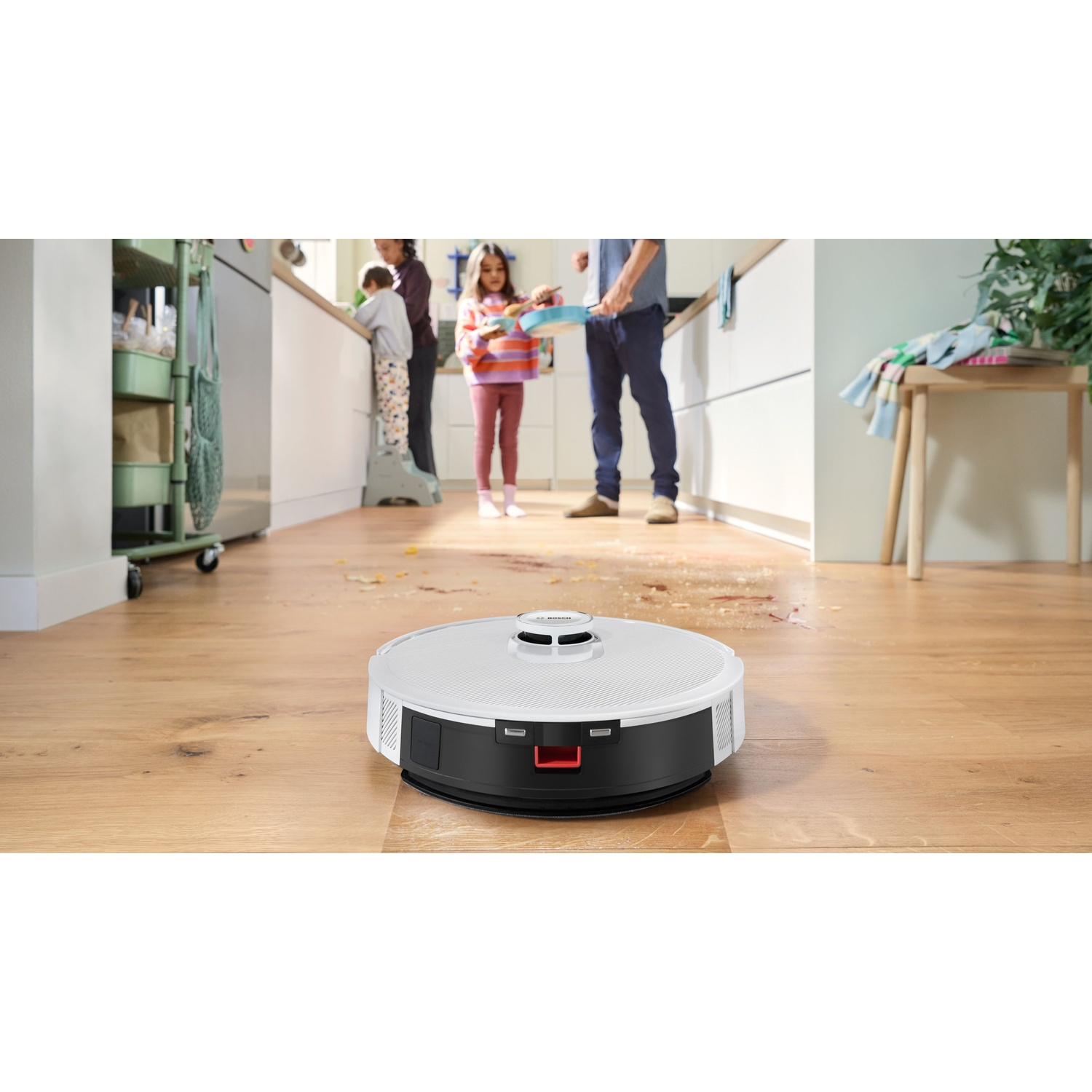 Un robot aspirapolvere su un pavimento in legno. Sullo sfondo, una famiglia è in cucina e c'è sporco sul pavimento.