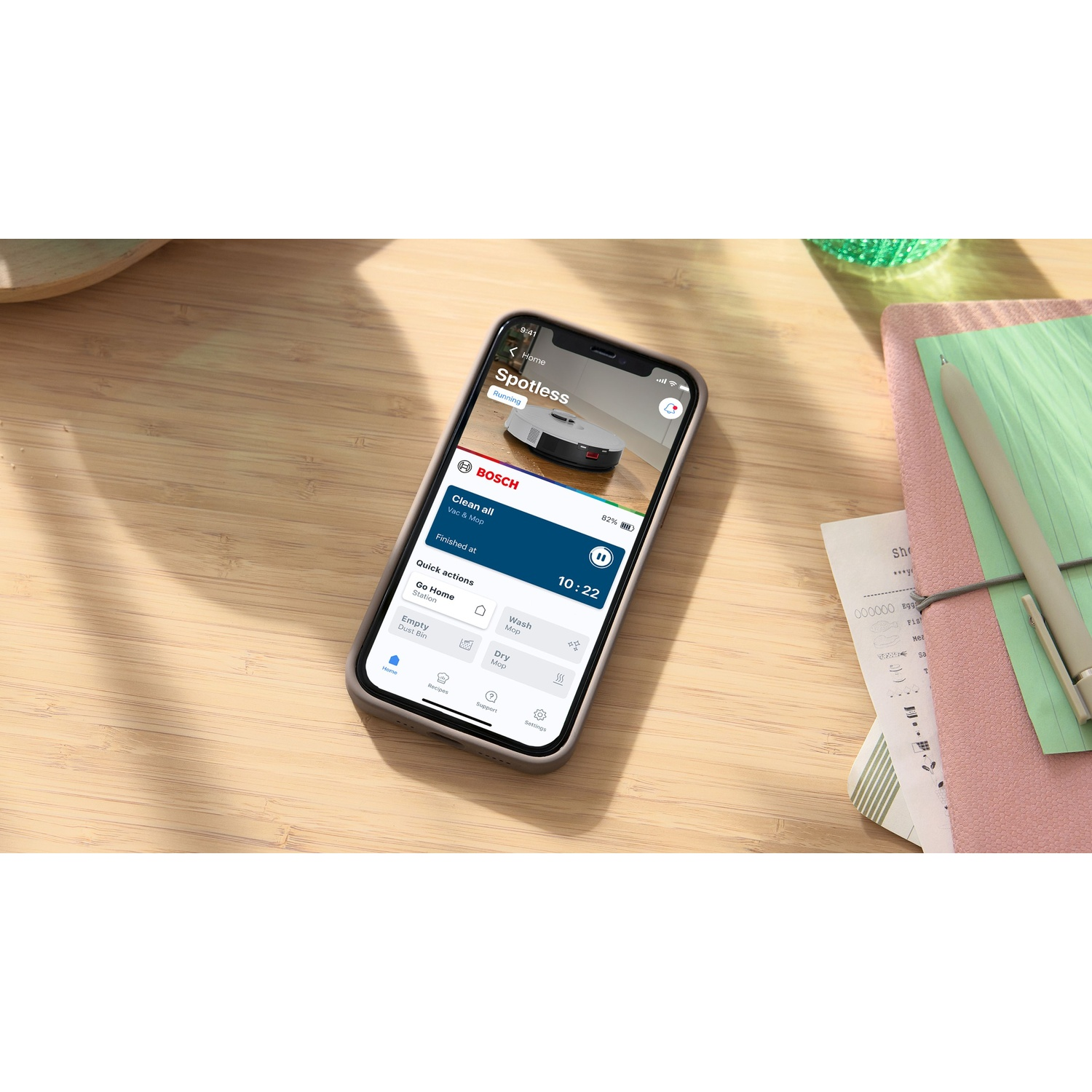 Uno smartphone mostra l'app Bosch Spotless. Mostra un robot aspirapolvere, un tavolo di legno, una penna e alcune ricevute.