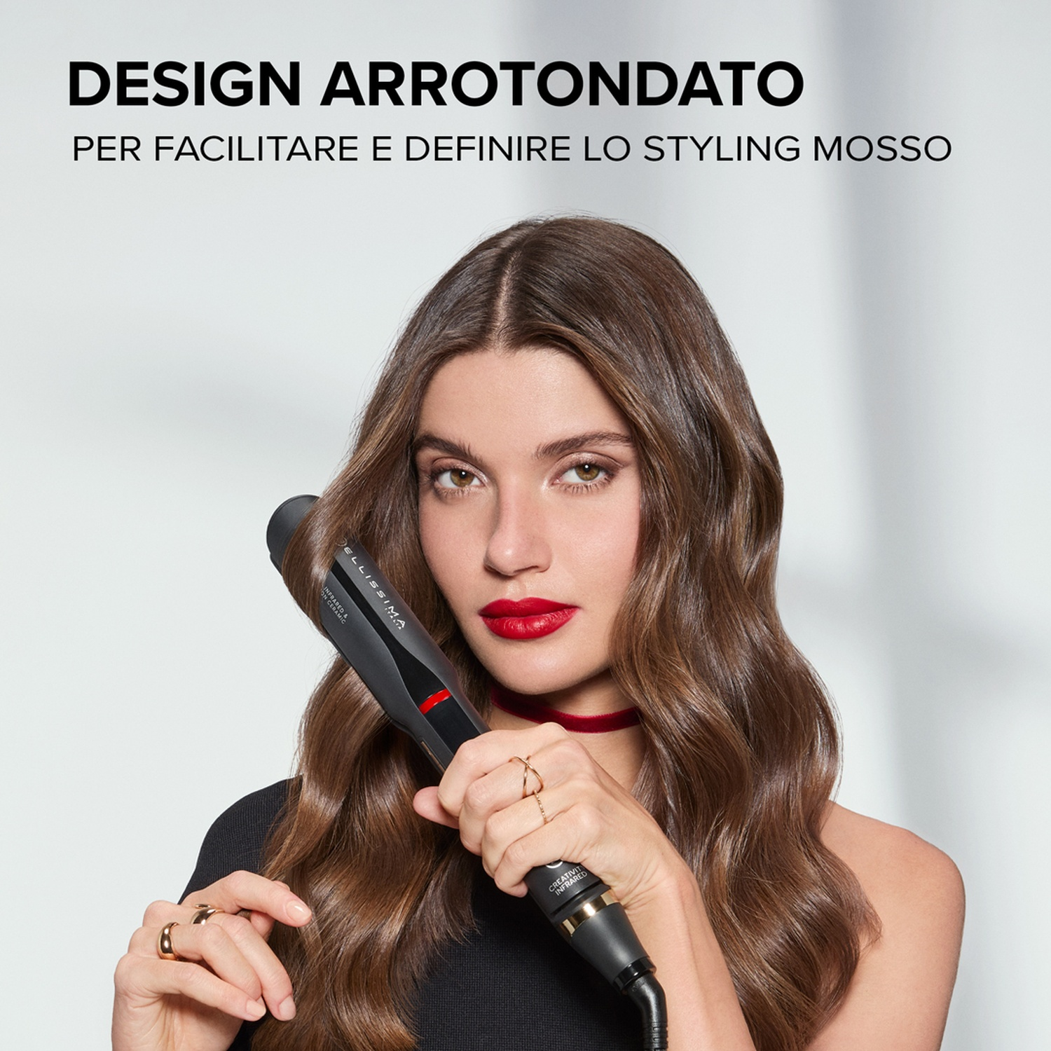Una donna con i capelli castani ondulati tiene in mano una piastra. Ha il rossetto rosso e un choker rosso. Lo sfondo è bianco.