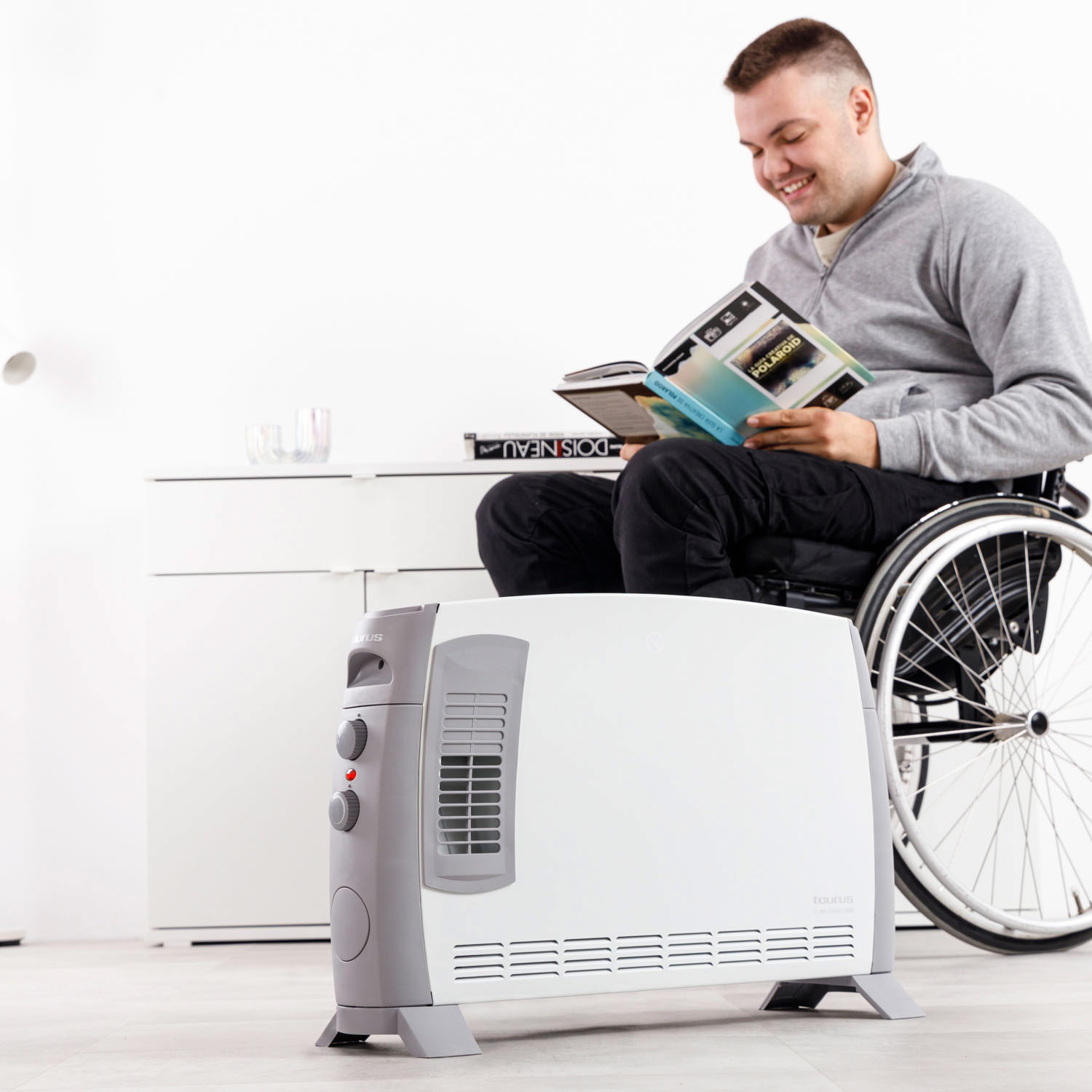 Hombre en silla de ruedas lee un libro, sonriendo. Un calefactor blanco está al frente. Fondo incluye un armario blanco y pared.