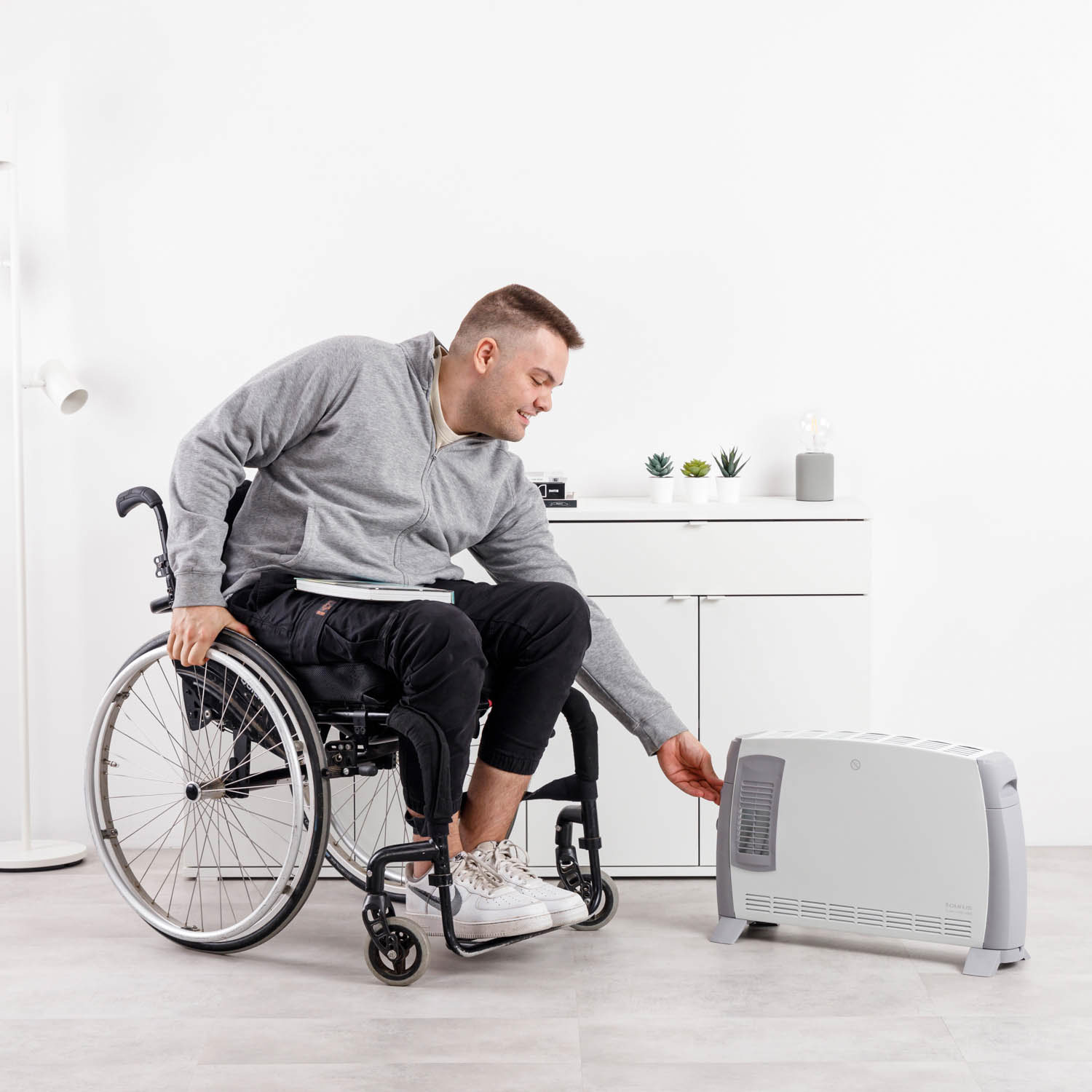Hombre en silla de ruedas se acerca a un calentador gris. Gabinete blanco y plantas en el fondo. Habitación luminosa con lámpara.