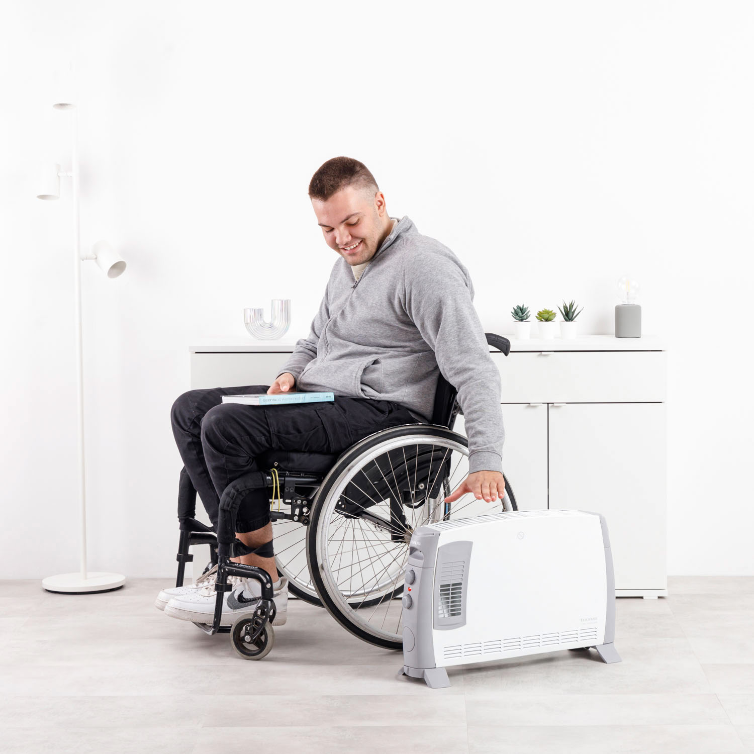 Un hombre en silla de ruedas sonríe. Sostiene un libro y alcanza un calefactor blanco. Un armario y una lámpara blancos están al fondo.