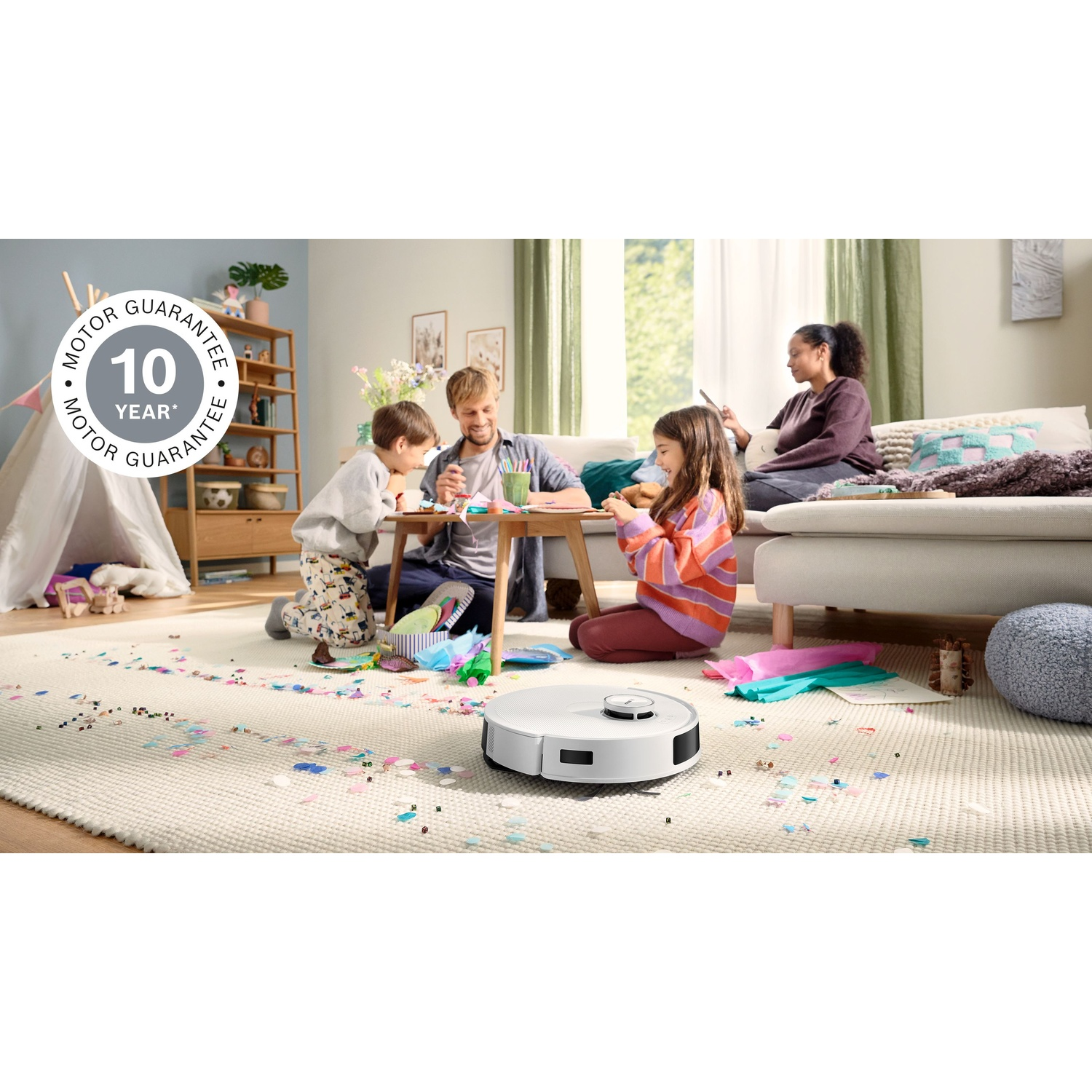Famiglia in soggiorno con un aspirapolvere robot. Bambini e genitori che creano. Coriandoli e una tenda sul pavimento. Una donna usa un tablet.