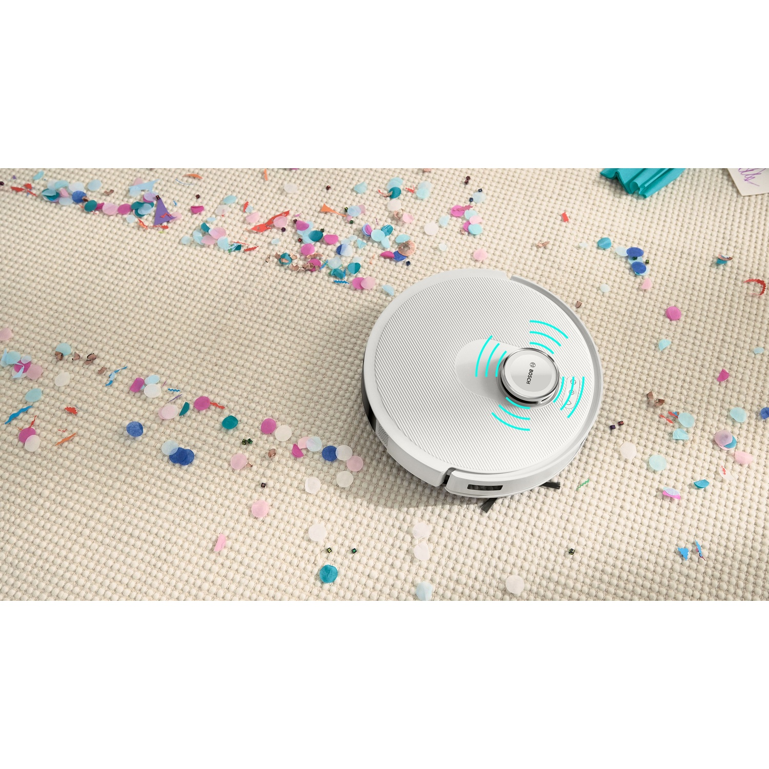 Un robot aspirapolvere bianco su un tappeto beige cosparso di coriandoli colorati. Il robot ha un anello illuminato blu.