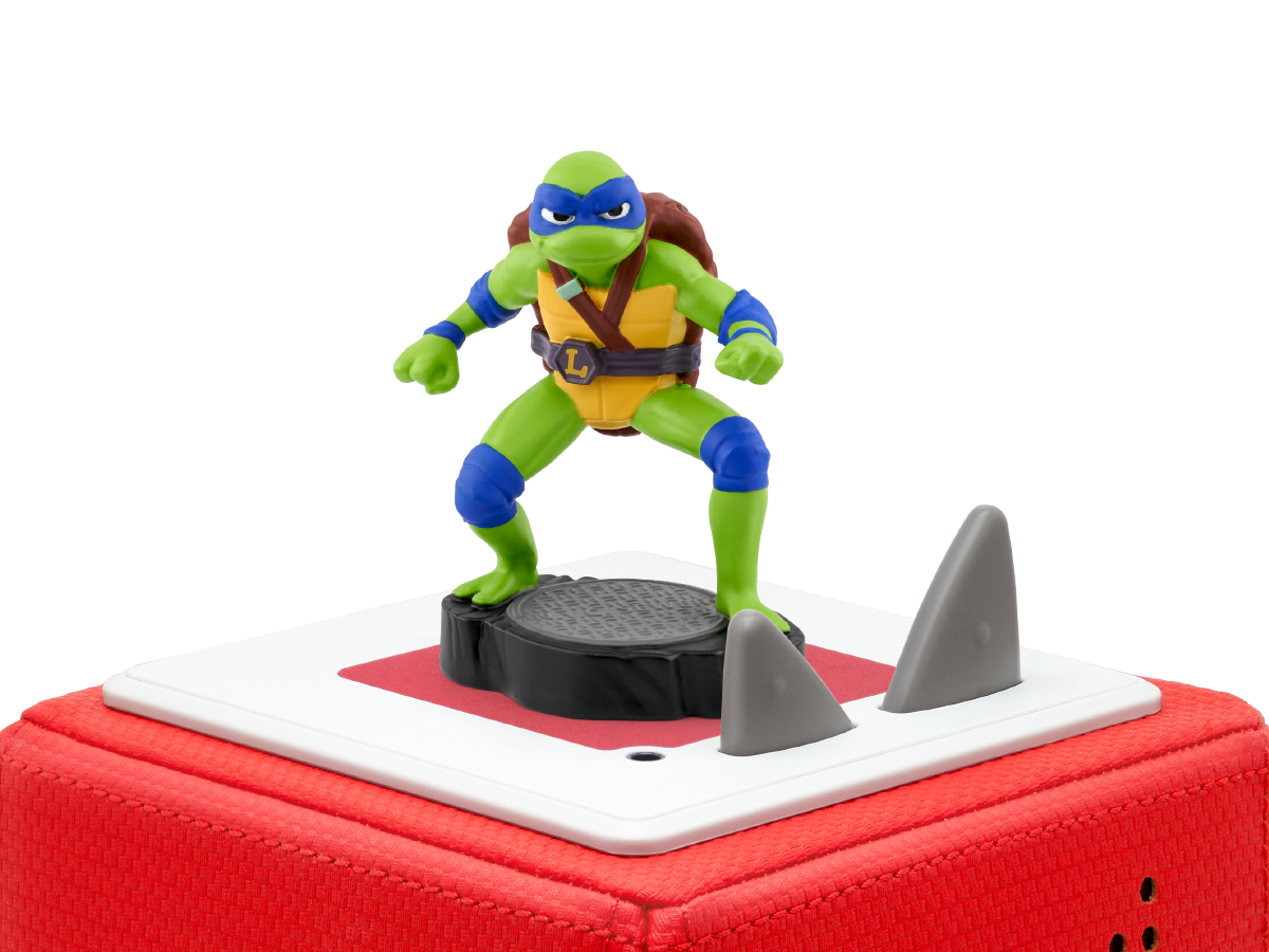 Eine Teenage Mutant Ninja Turtle-Figur steht auf einer Plattform mit Haifischflossen, die sich auf einem roten Würfel befindet. Die Schildkröte ist grün, blau und gelb.