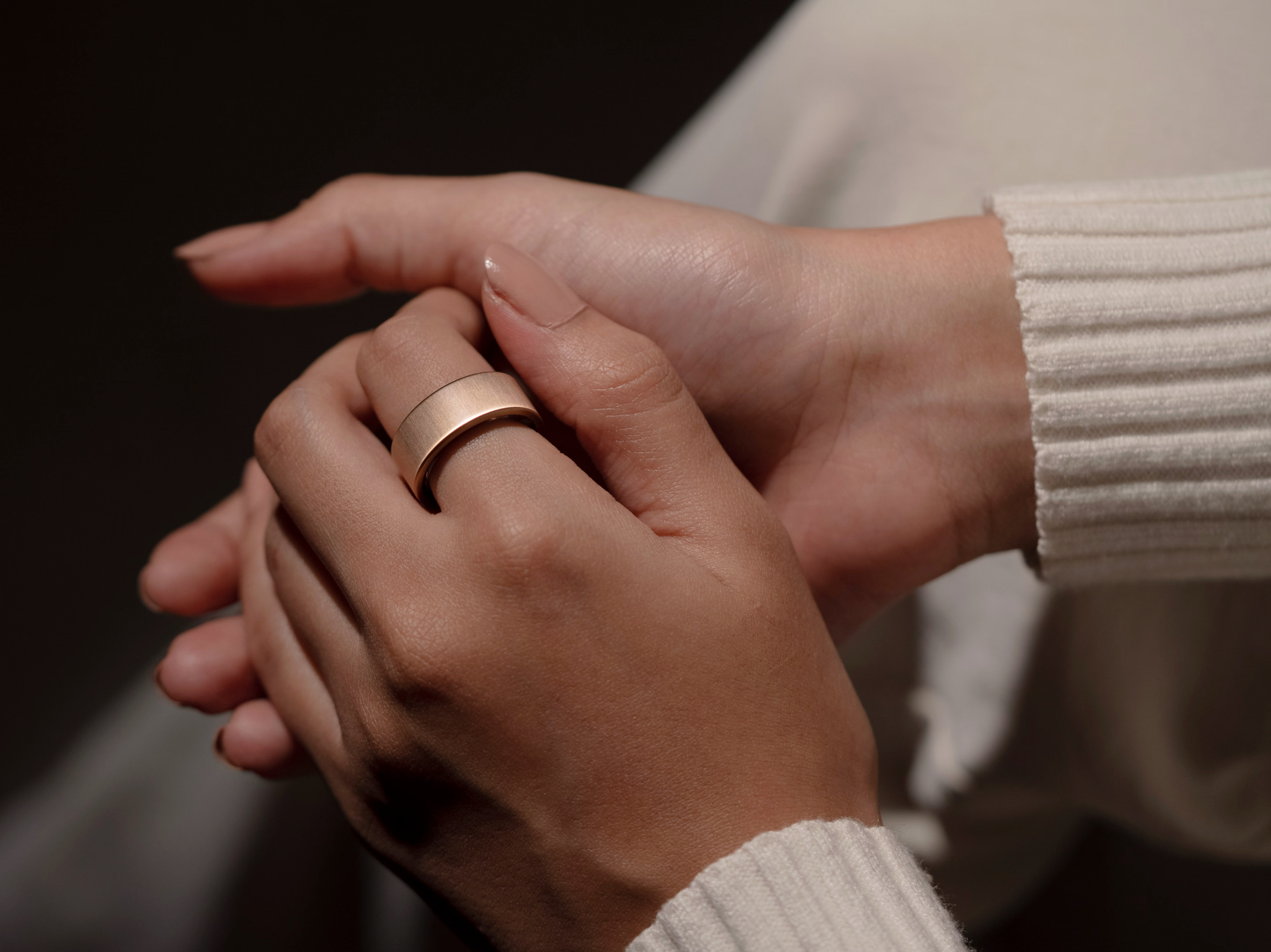 Dłoń z złotym pierścieniem, spoczywająca na innej dłoni. Obie dłonie na kremowym swetrze. Ciemne tło.
