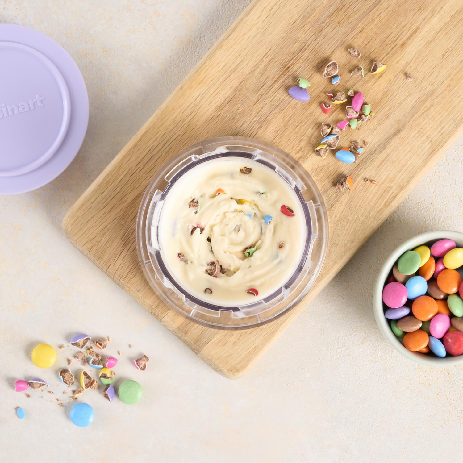 Draufsicht auf Eis mit Süßigkeiten-Toppings, Holzbrett, lila Deckel und eine Schüssel mit bunten Süßigkeiten auf hellem Hintergrund.