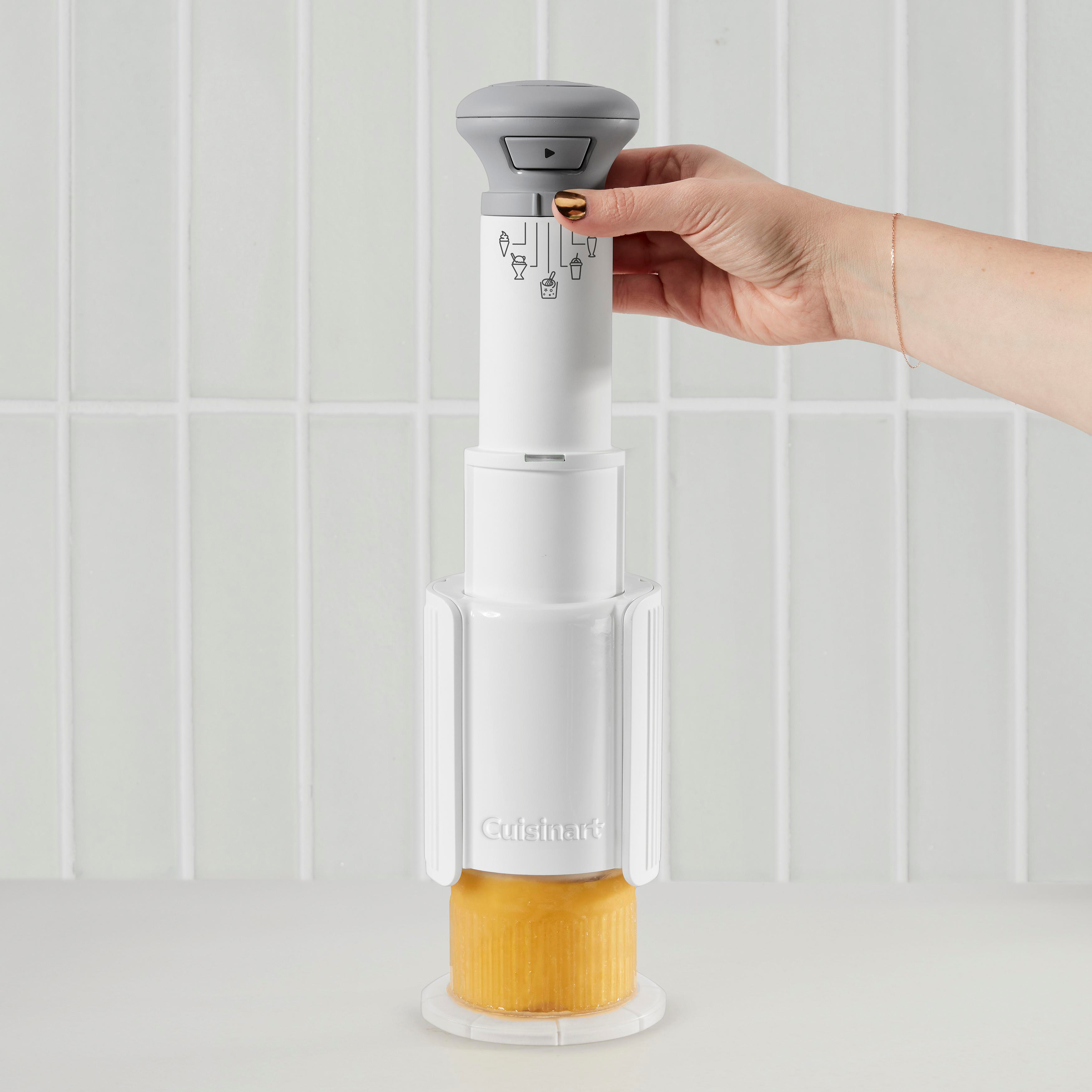 Eine Hand hält eine weiße Cuisinart-Eismaschine über einem gelben Eisblock. Der Hintergrund ist eine weiße Fliesenwand.