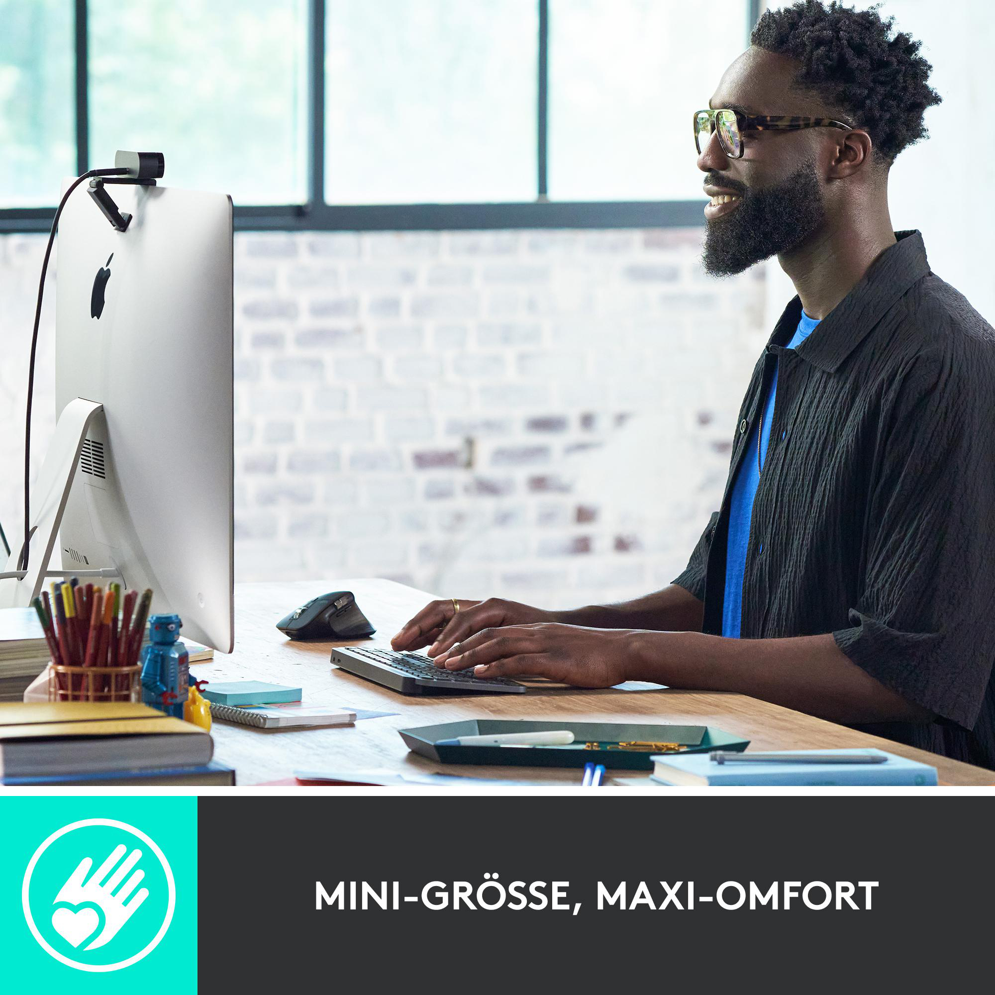 Ein Mann mit Brille tippt auf einer Tastatur. Ein Monitor und eine Webcam sind vorhanden. Der Hintergrund ist eine Ziegelmauer.