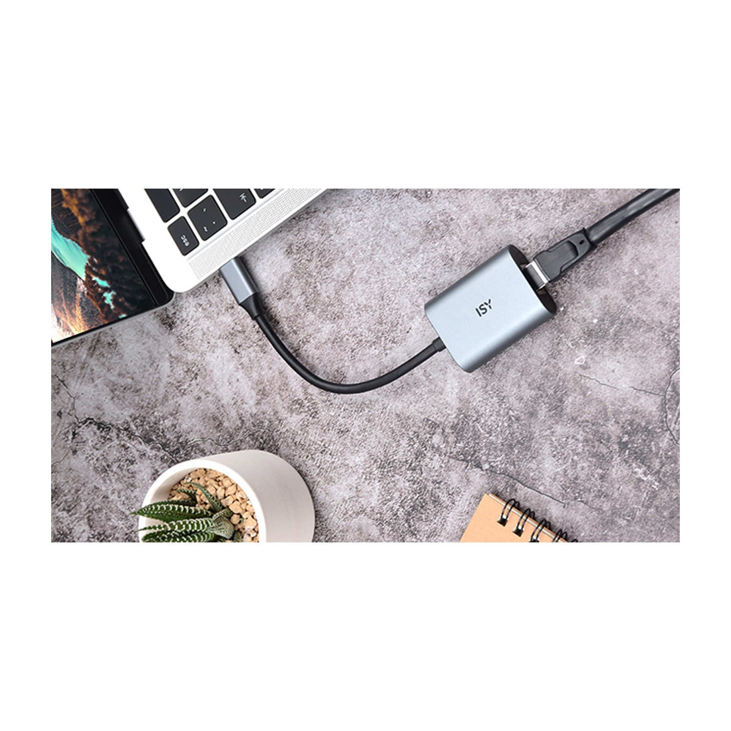 Un laptop collegato a un adattatore argento con l'etichetta 'ISY'. Una pianta e un taccuino vicino al laptop su una superficie strutturata.