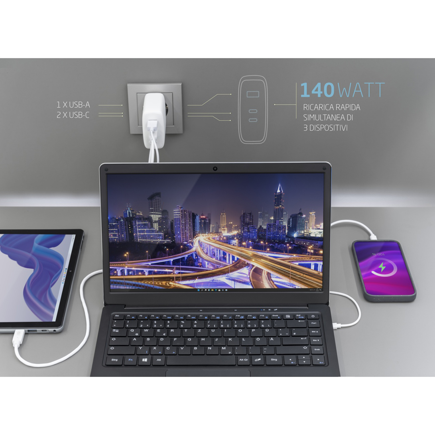 Un laptop, un tablet e un telefono sono collegati e in carica. È visibile anche una presa a muro con un caricatore. Il laptop mostra una città.