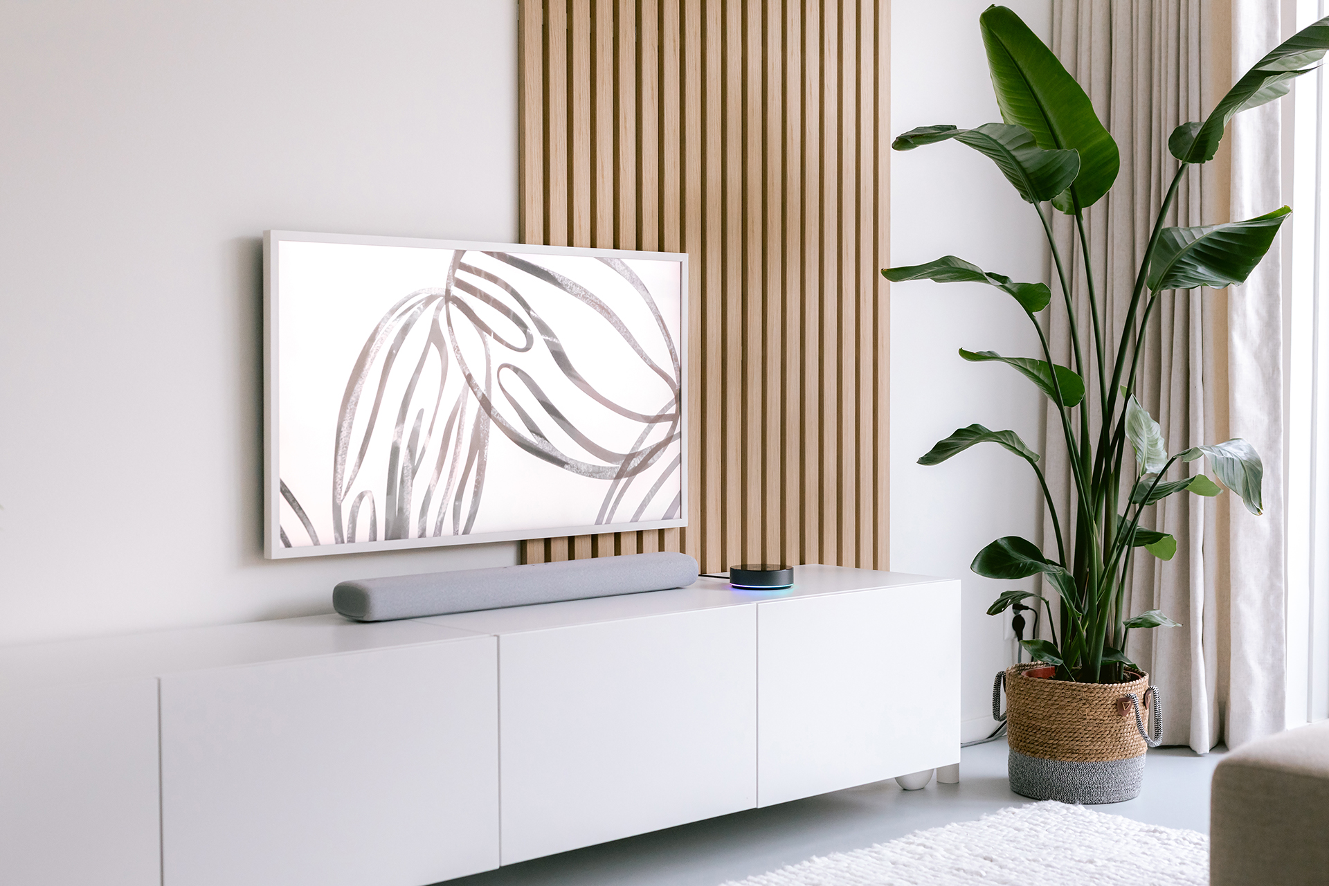 Salón con mueble blanco, TV, planta y panel de madera. El televisor muestra arte abstracto, una barra de sonido debajo.