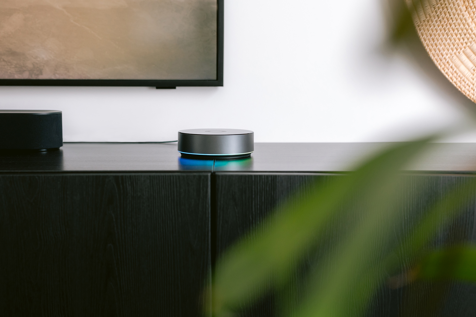 Un pequeño altavoz plateado con un anillo brillante azul está sobre un armario negro. Un televisor y una pared están en el fondo.