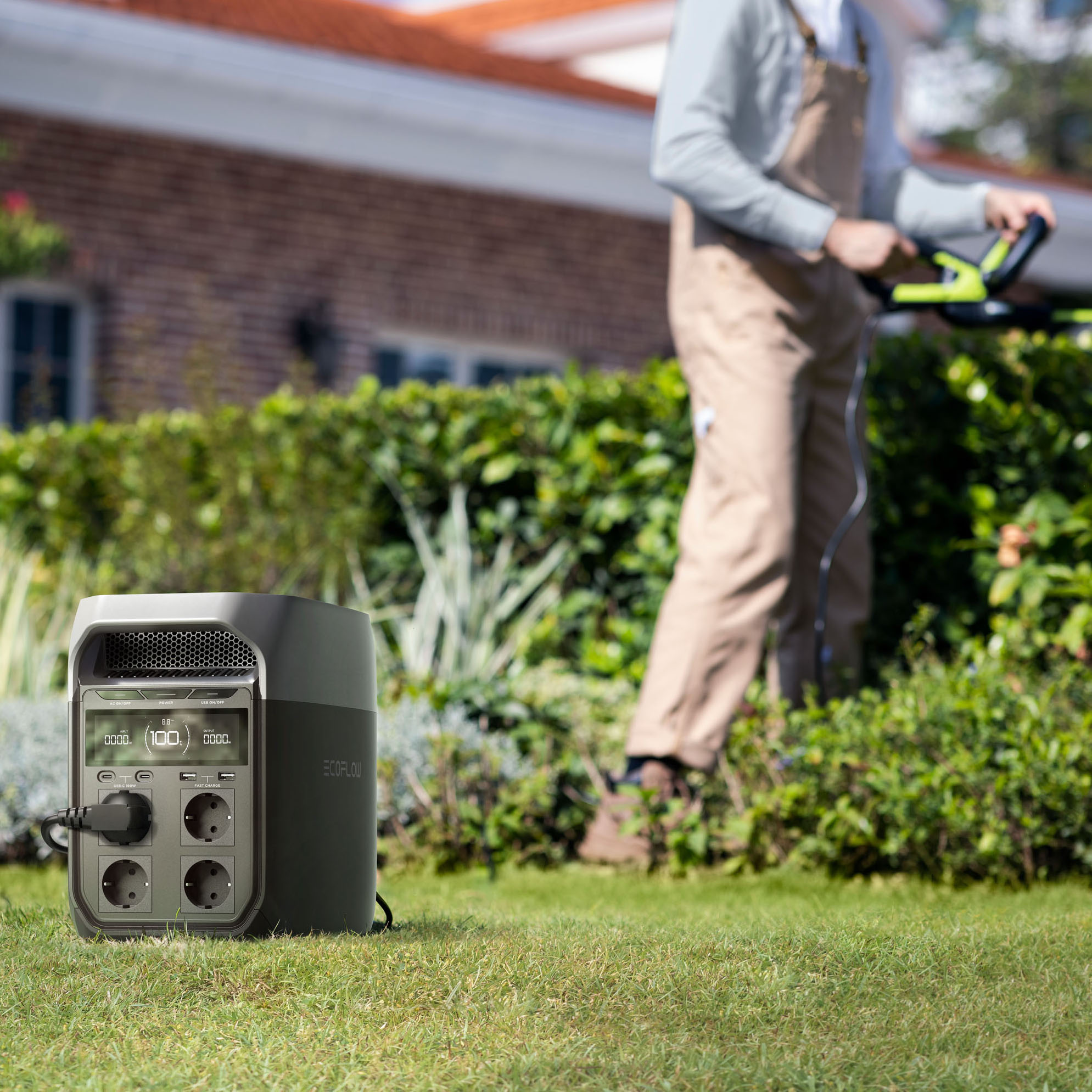 Una persona corta el césped con un cortacésped eléctrico, con una estación de energía portátil en primer plano. Casa al fondo.