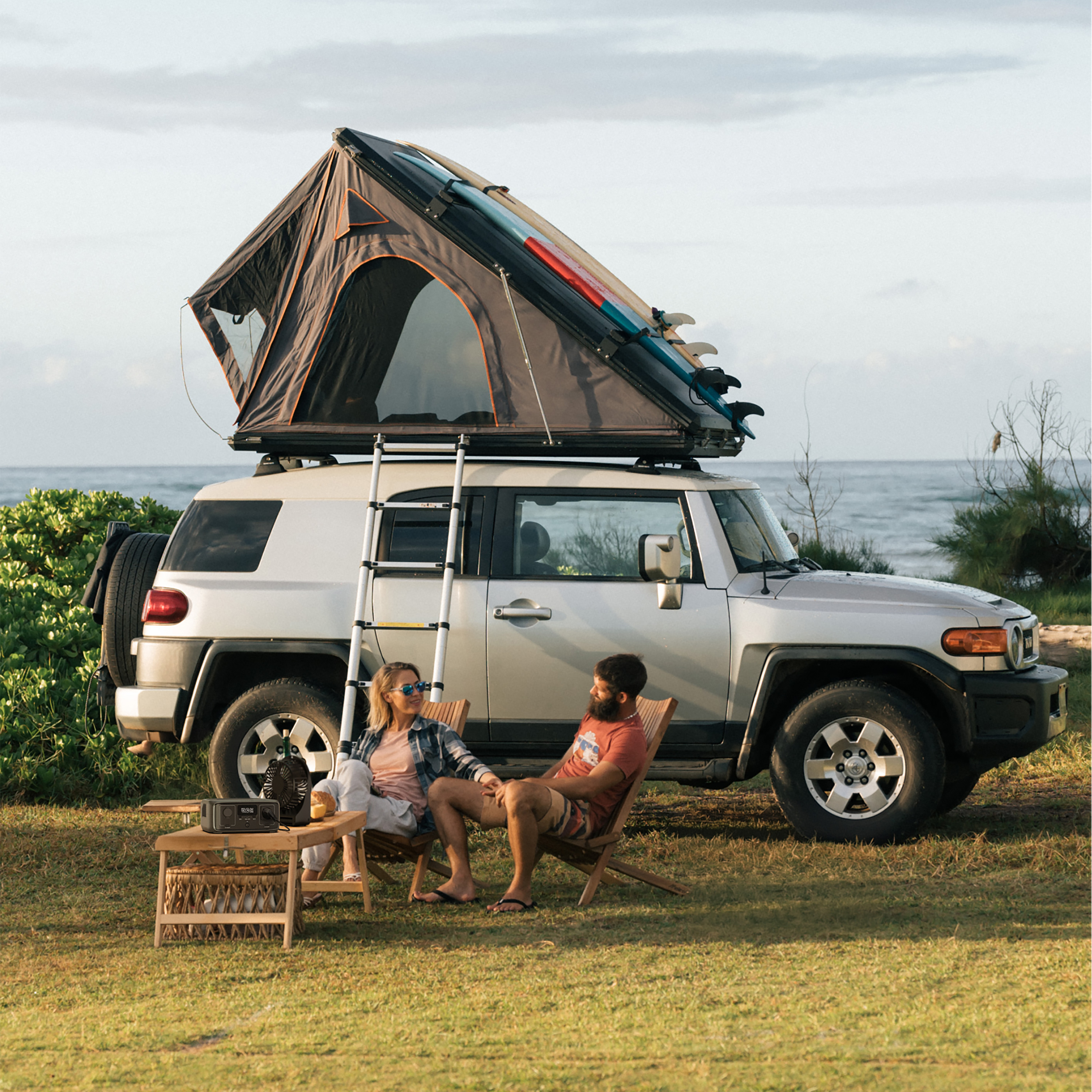 Una pareja se sienta cerca de un SUV plateado con tienda de techo, tablas de surf y una escalera. Playa y océano al fondo.