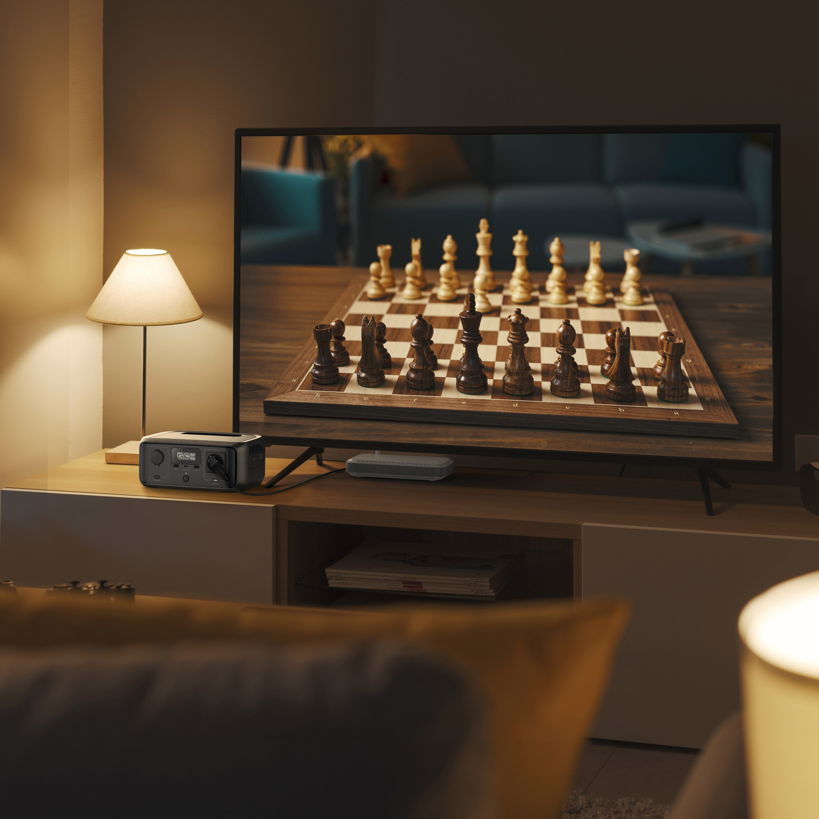 Un televisor muestra una partida de ajedrez. Hay una lámpara a la izquierda. Una estación de energía y un mando a distancia en el mueble.