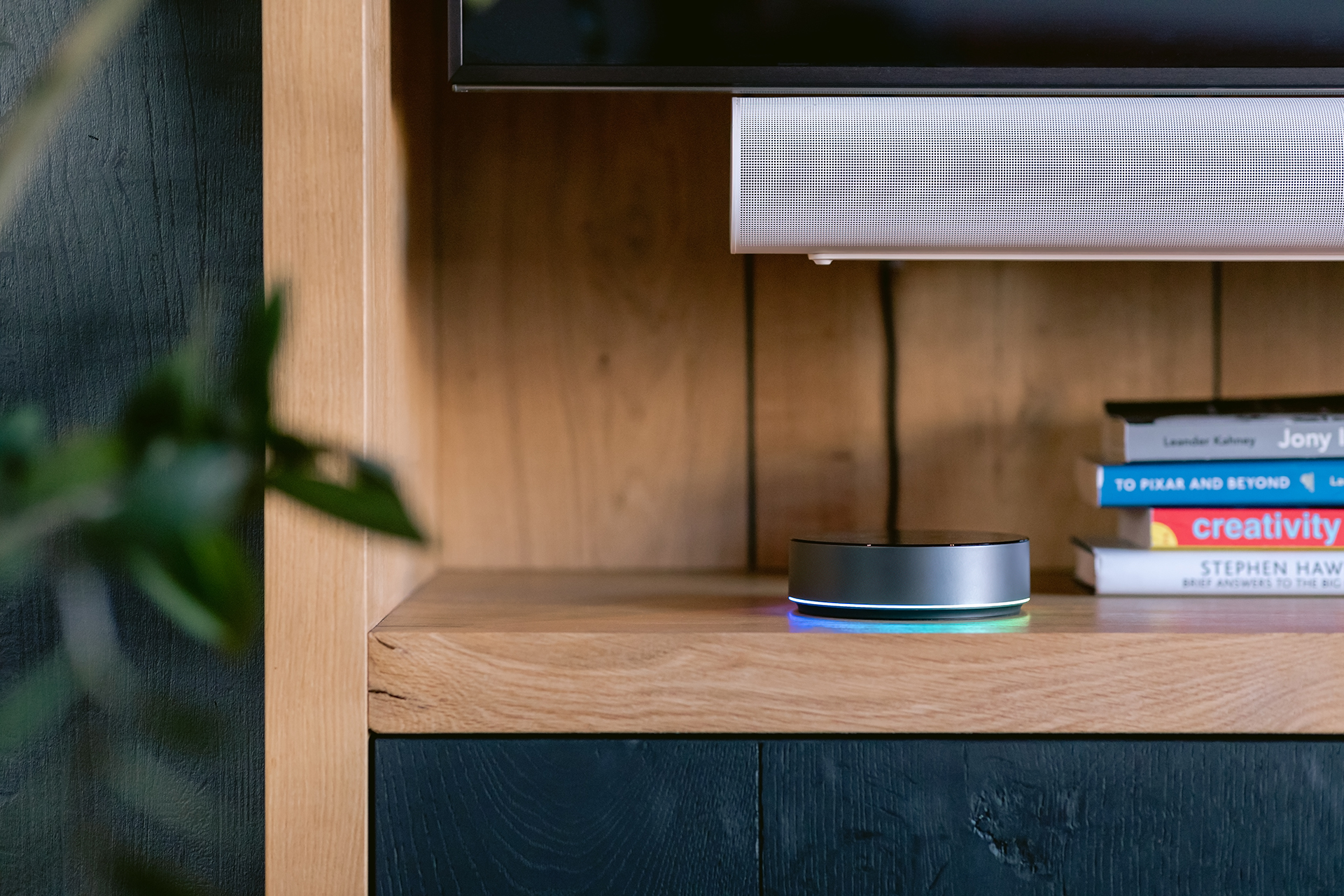 Un dispositivo con un anillo brillante se encuentra en un estante de madera con libros. Un altavoz y un televisor arriba. Planta verde en la esquina.