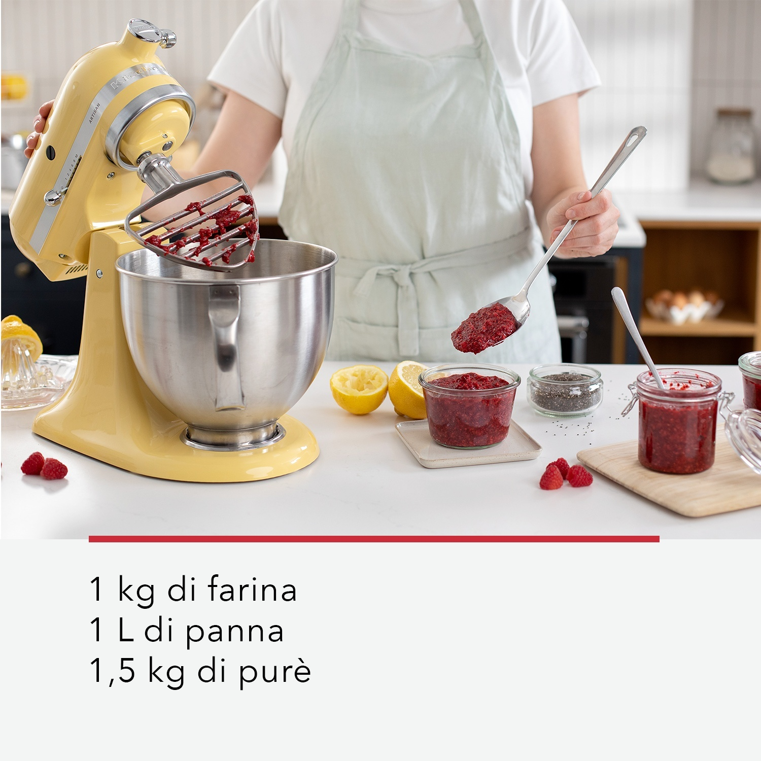 Una donna sta facendo la marmellata con un robot da cucina giallo. Gli ingredienti, tra cui lamponi e limoni, sono sul bancone. Sono visibili barattoli di marmellata.