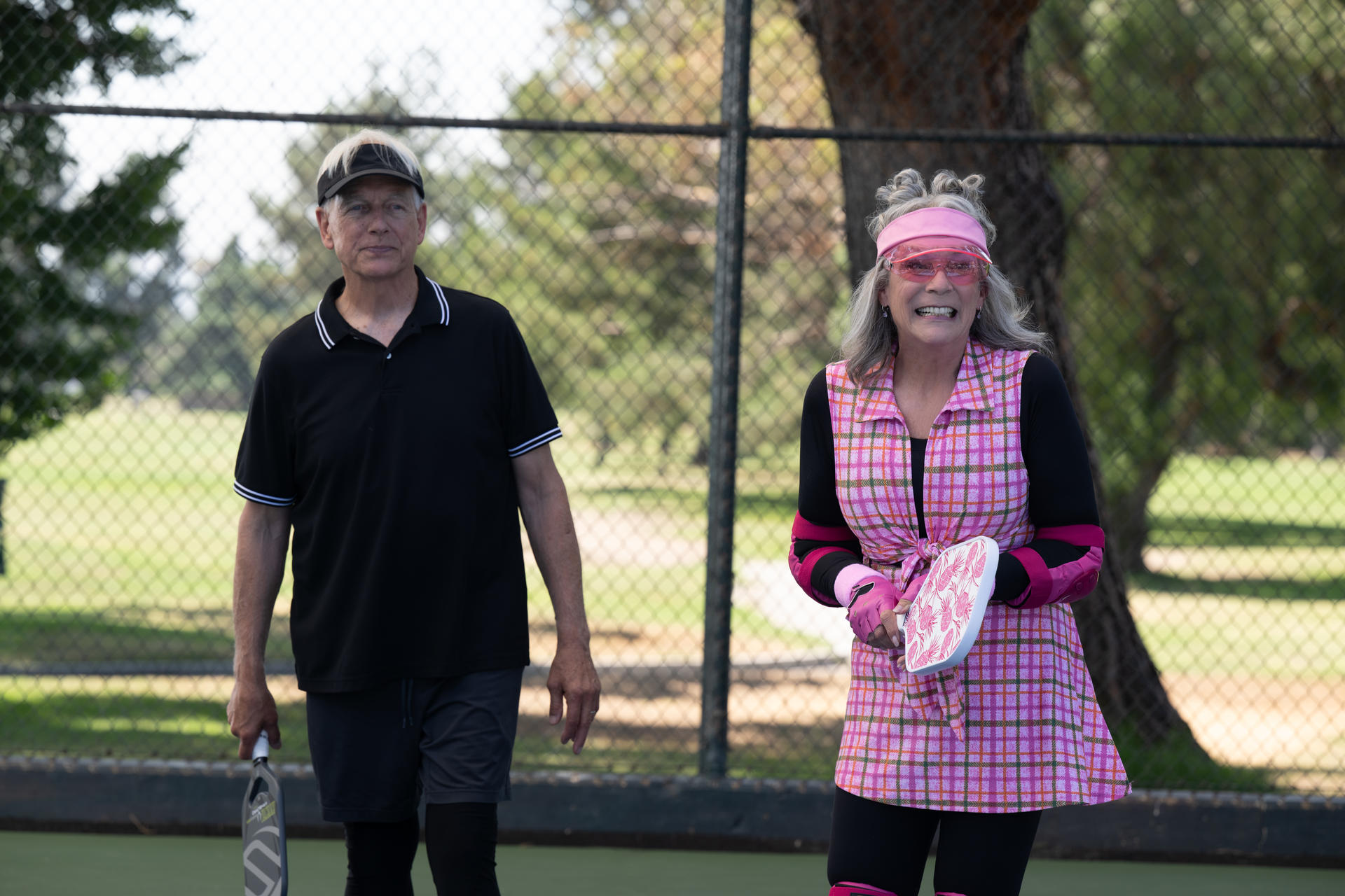 Ein Mann und eine Frau stehen auf einem Pickleball-Platz. Die Frau trägt rosa Karos und ein Visier, der Mann ein schwarzes Hemd.