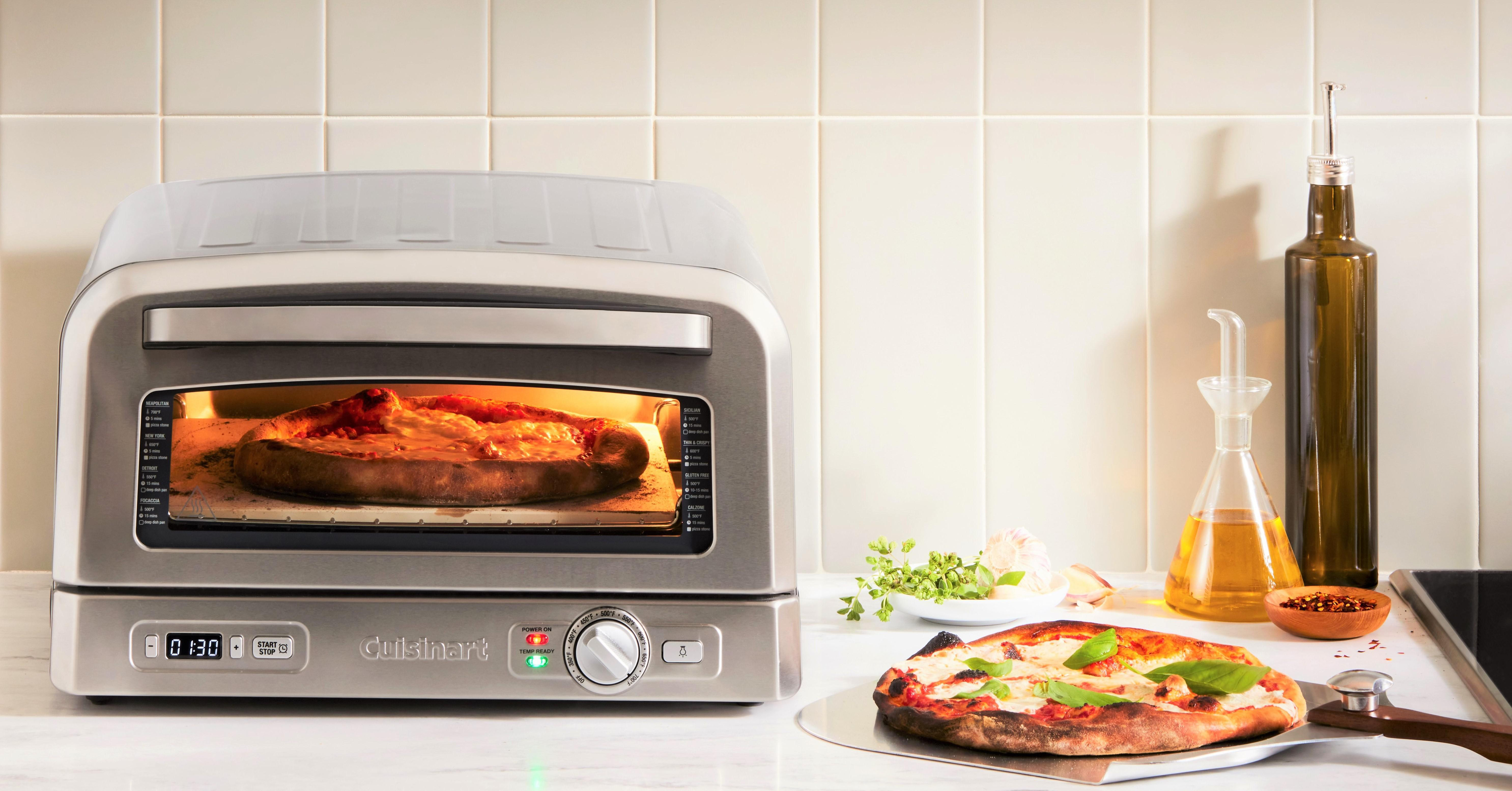 Ein silberner Pizzaofen mit einer Pizza darin, eine weitere Pizza auf einem Schieber und Olivenölflaschen.