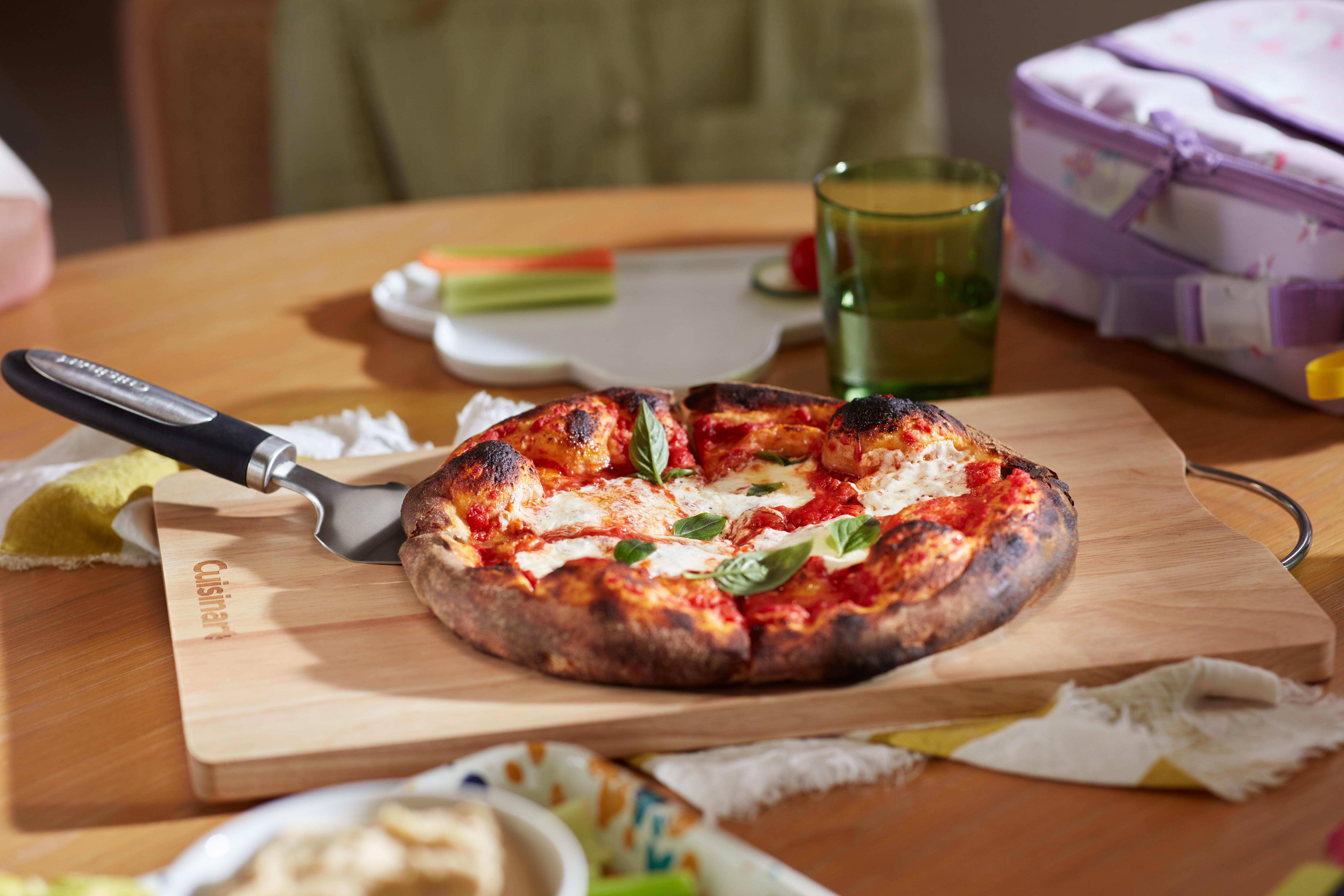 Eine Pizza mit Basilikum auf einem Holzbrett, ein Glas grüne Flüssigkeit und eine Brotdose stehen auf einem Holztisch.