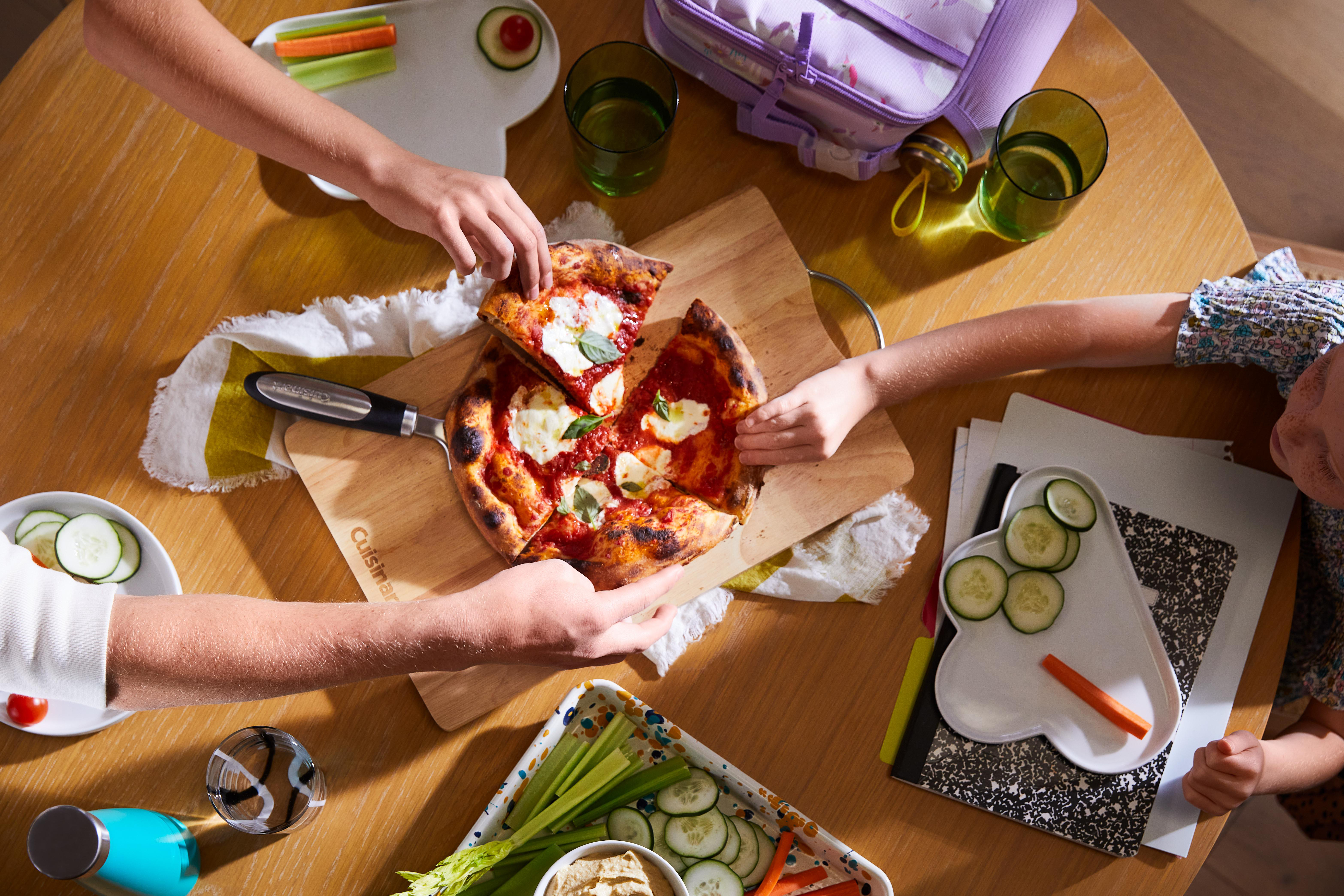 Ein runder Holztisch, an dem Menschen nach Pizza und Snacks greifen. Pizza, Gemüse und Getränke stehen auf dem Tisch.