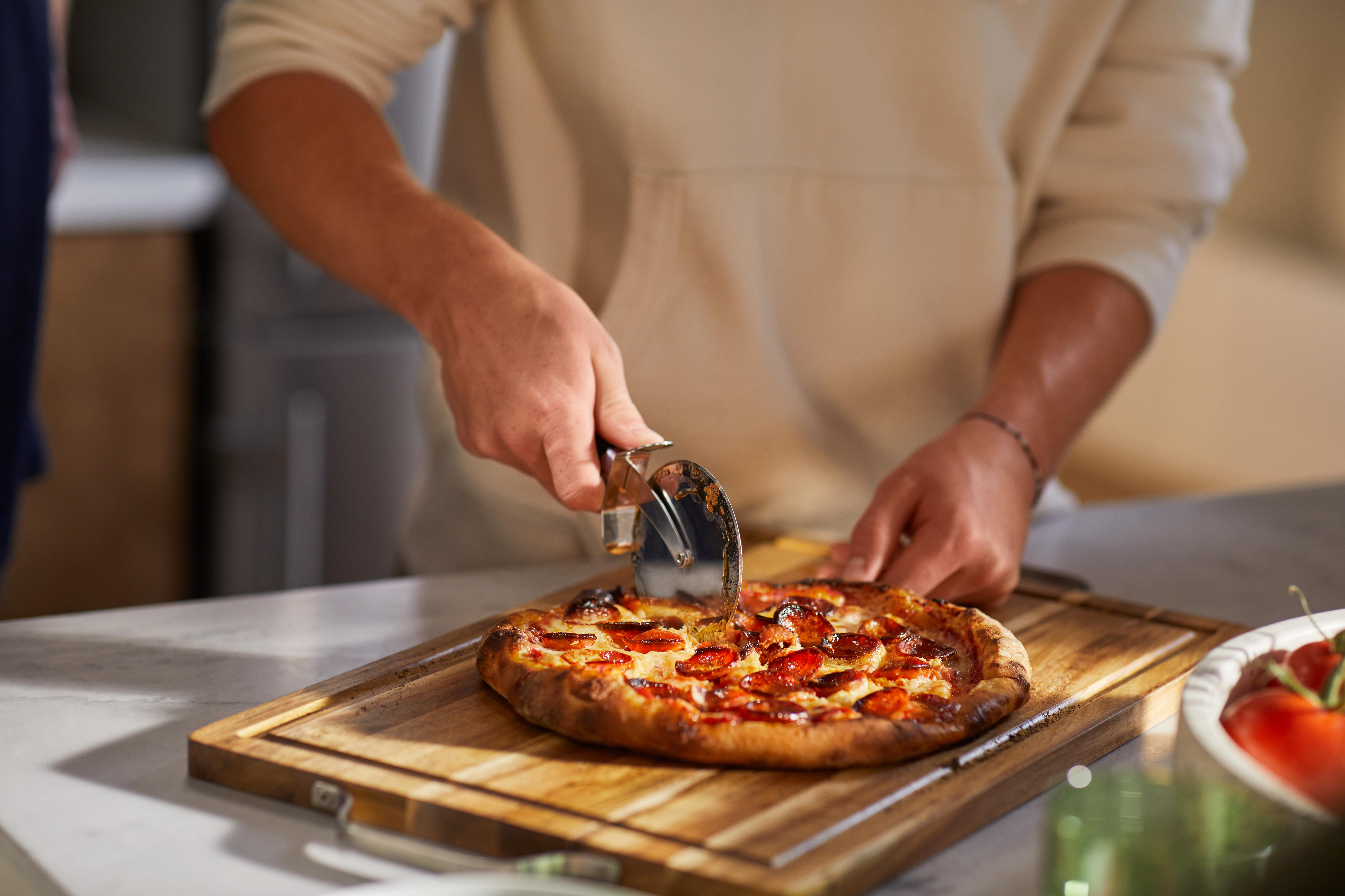 Eine Person schneidet eine Peperoni-Pizza auf einem Holzbrett mit einem Pizzaschneider. Die Pizza liegt auf einer Marmortheke.