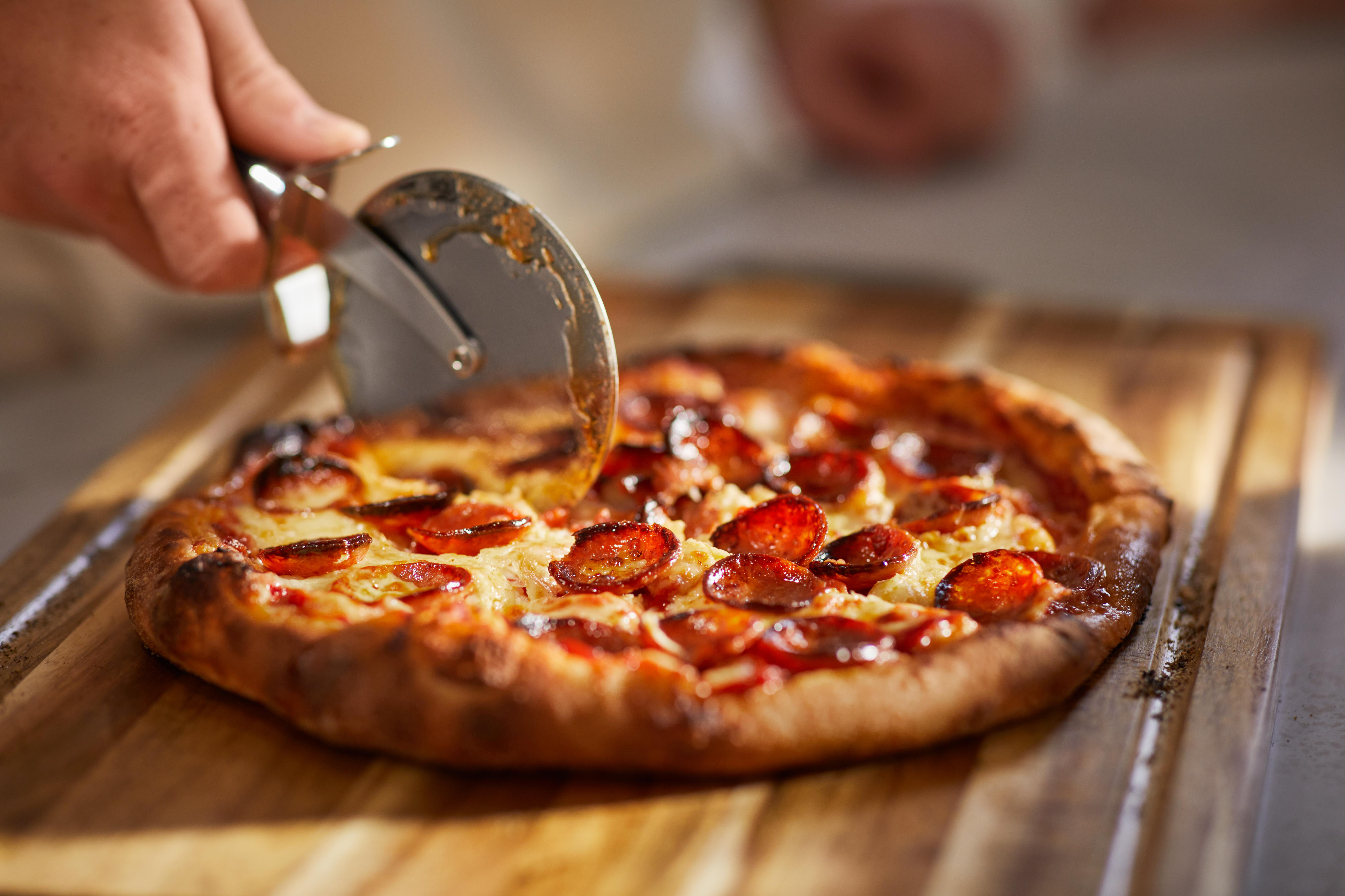 Eine Hand schneidet eine Peperoni-Pizza mit einem Pizzaschneider auf einem Holzbrett. Die Pizza ist rund und hat Käse.