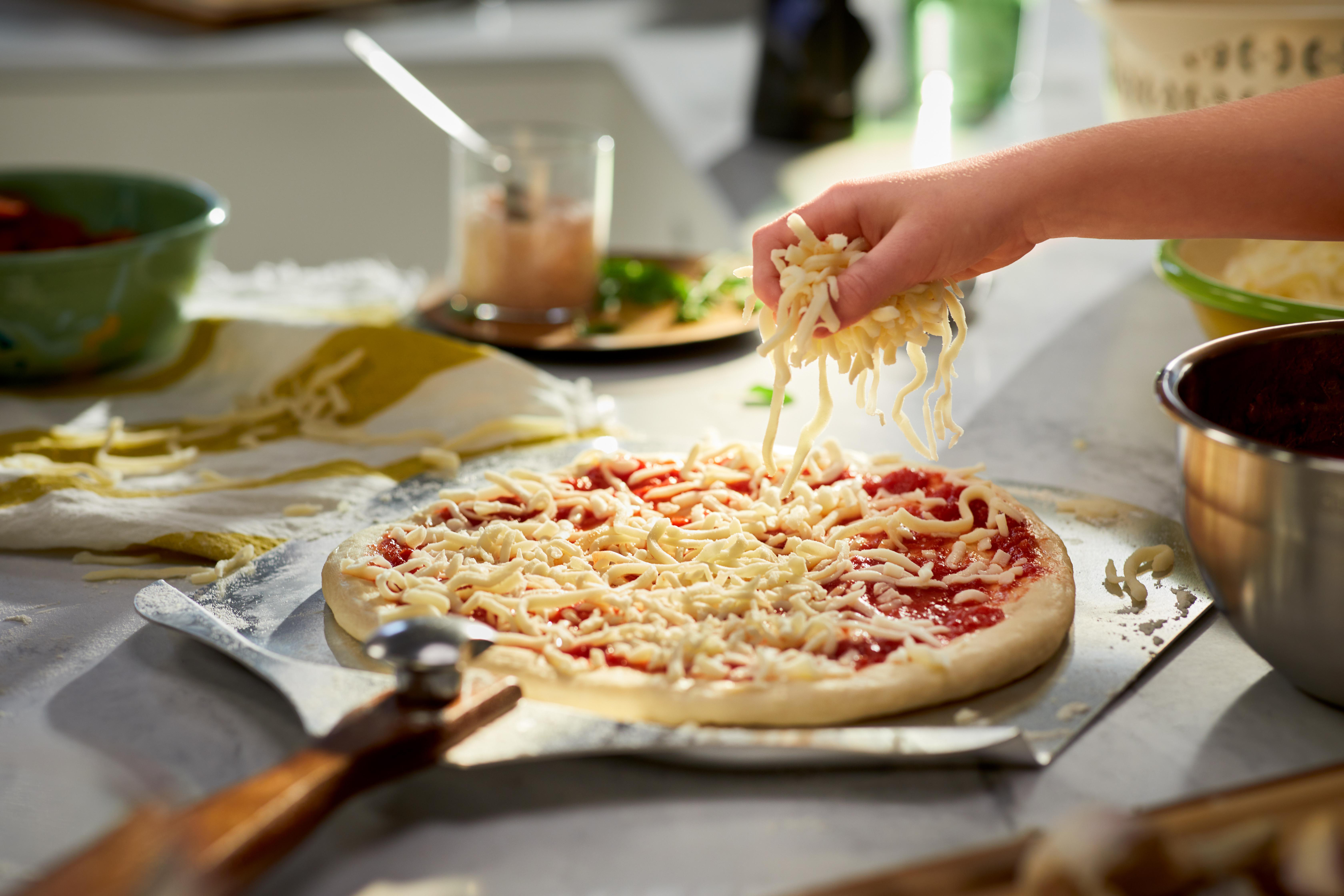 Eine Hand streut geriebenen Käse auf eine Pizza mit Tomatensauce. Andere Zutaten und Utensilien sind im Hintergrund.