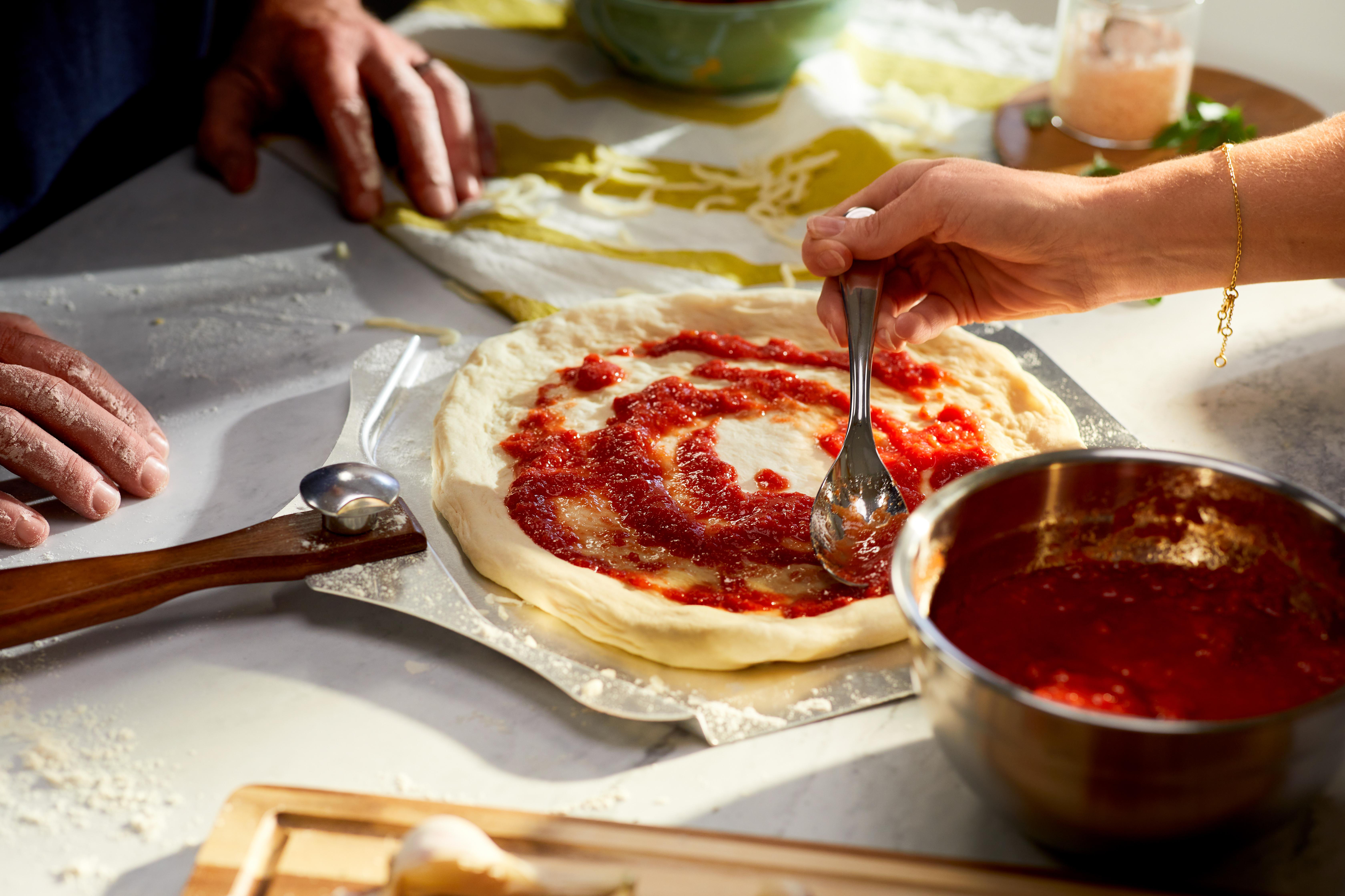Eine Person verteilt Tomatensauce mit einem Löffel auf Pizzateig. Hände, Teig, Tomatensauce und Schüsseln sind zu sehen.