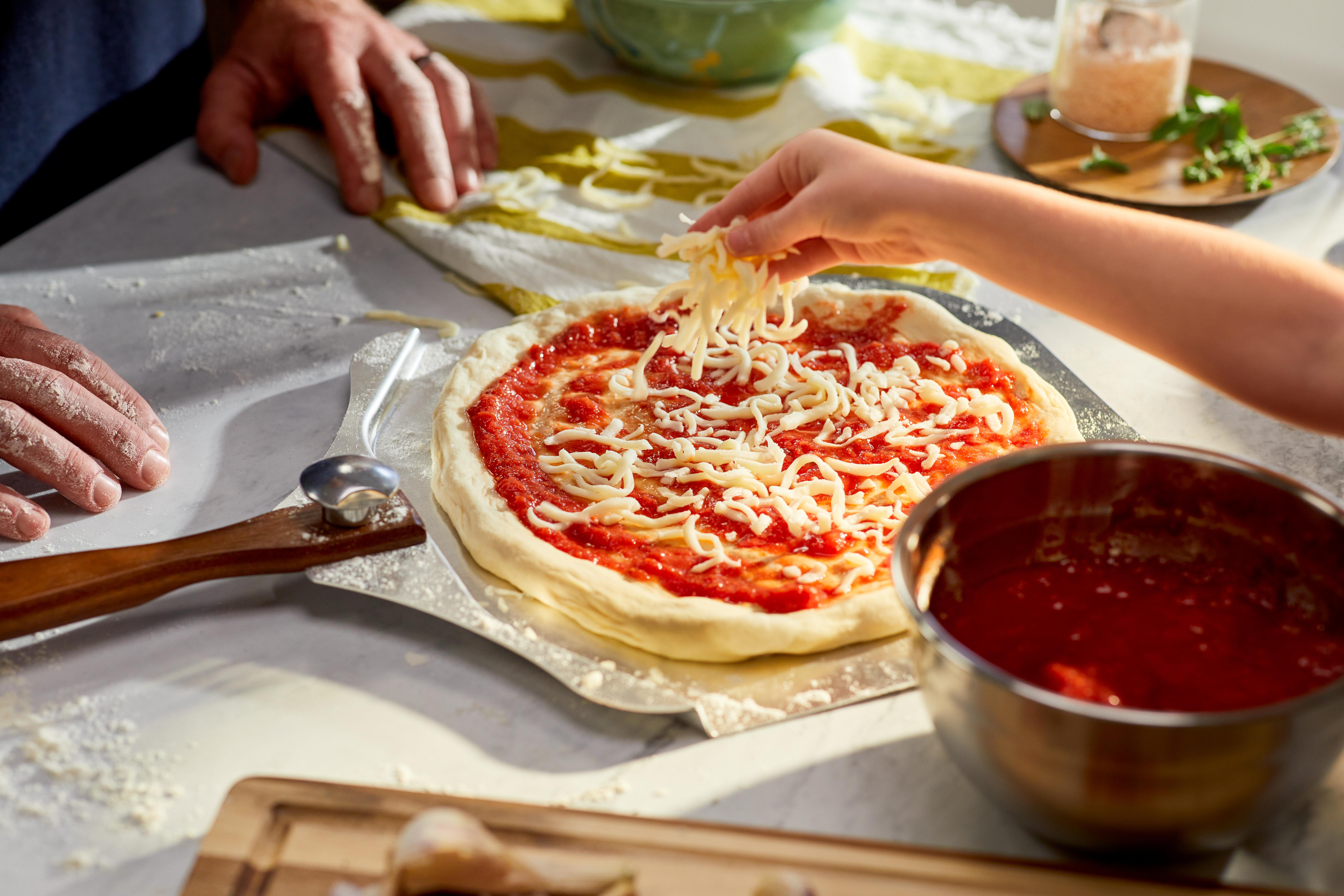 Eine Pizza wird zubereitet. Eine Hand streut Käse über die Tomatensauce. Weitere Zutaten sind sichtbar.