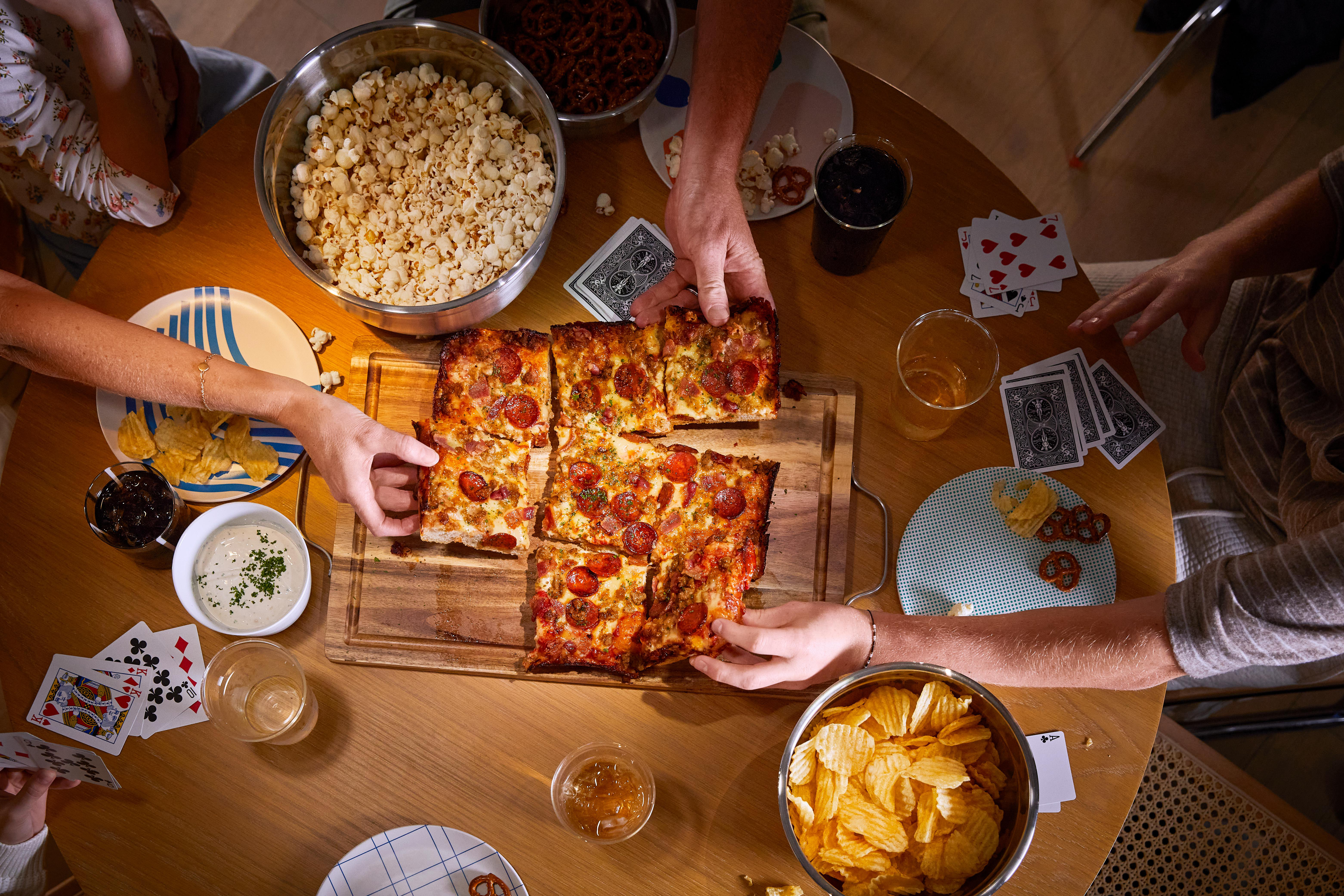 Ein runder Tisch mit Pizza, Popcorn, Chips, Brezeln und Spielkarten. Mehrere Hände greifen nach Essen.