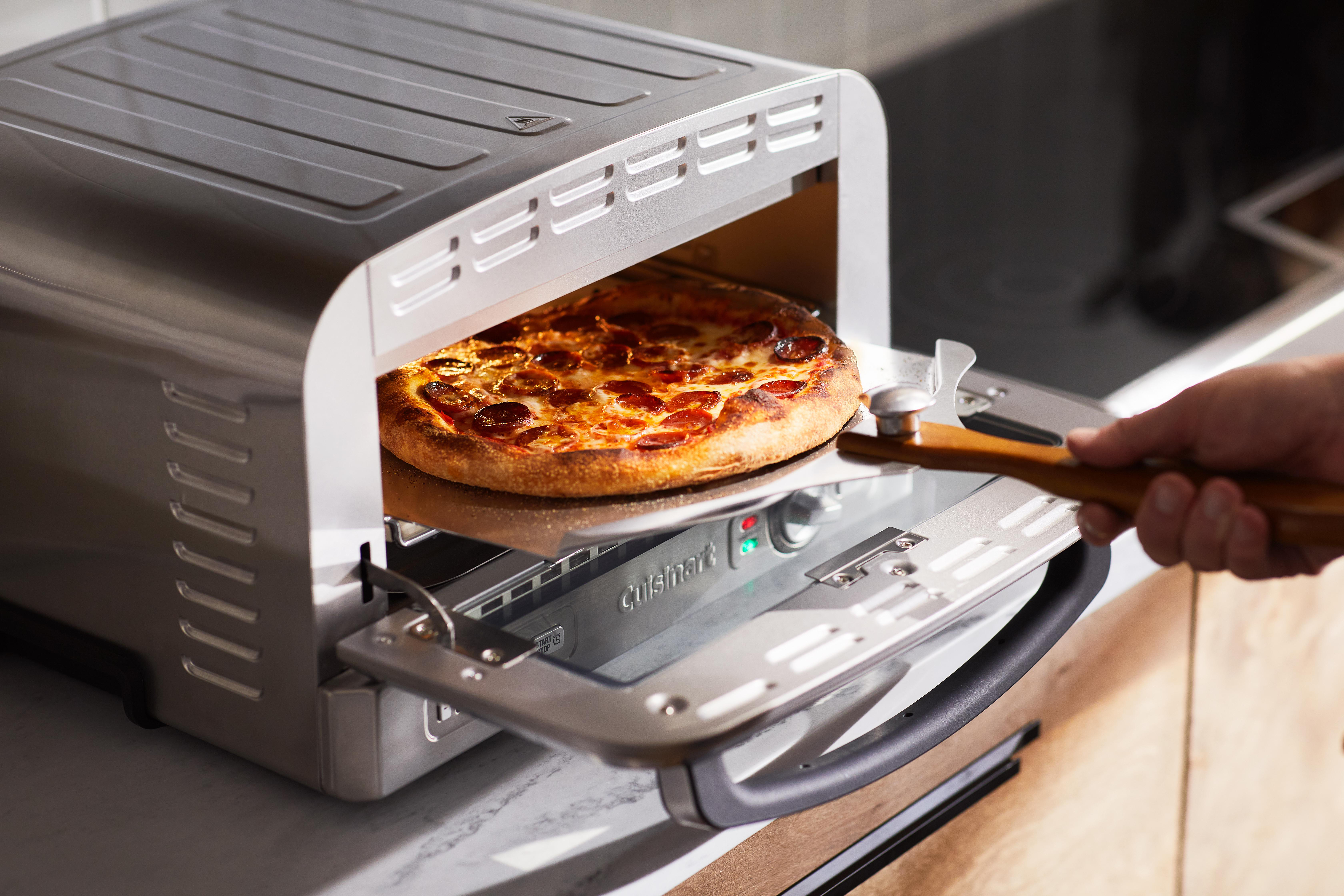 Eine Pizza wird aus einem Cuisinart-Pizzaofen genommen. Eine Hand benutzt einen Holzschieber, um die Pizza anzuheben.