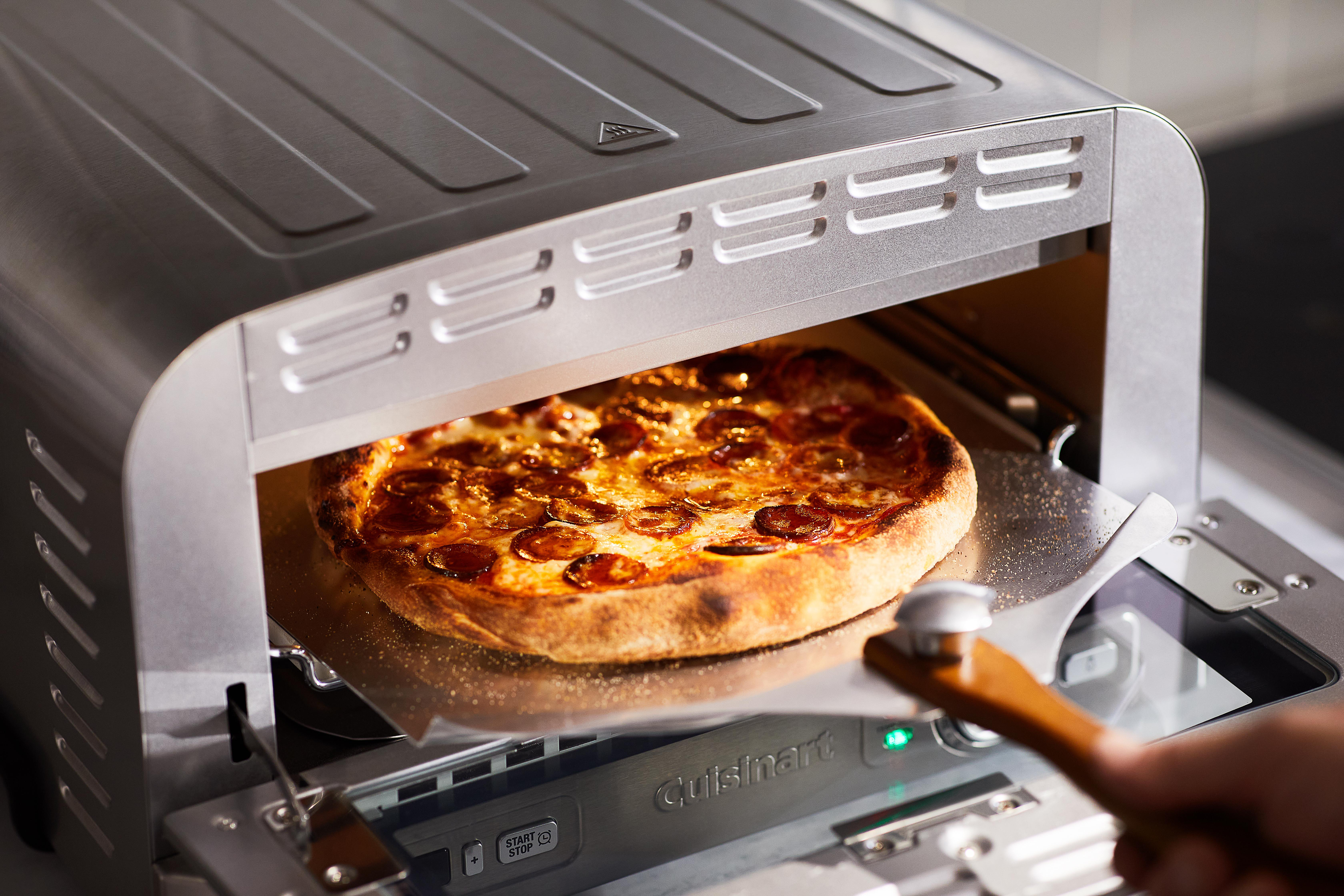 Eine Pepperoni-Pizza wird in einem Cuisinart-Ofen gebacken. Ein hölzerner Pizzaschieber greift hinein. Der Ofen ist silberfarben. Die Pizza ist goldbraun.