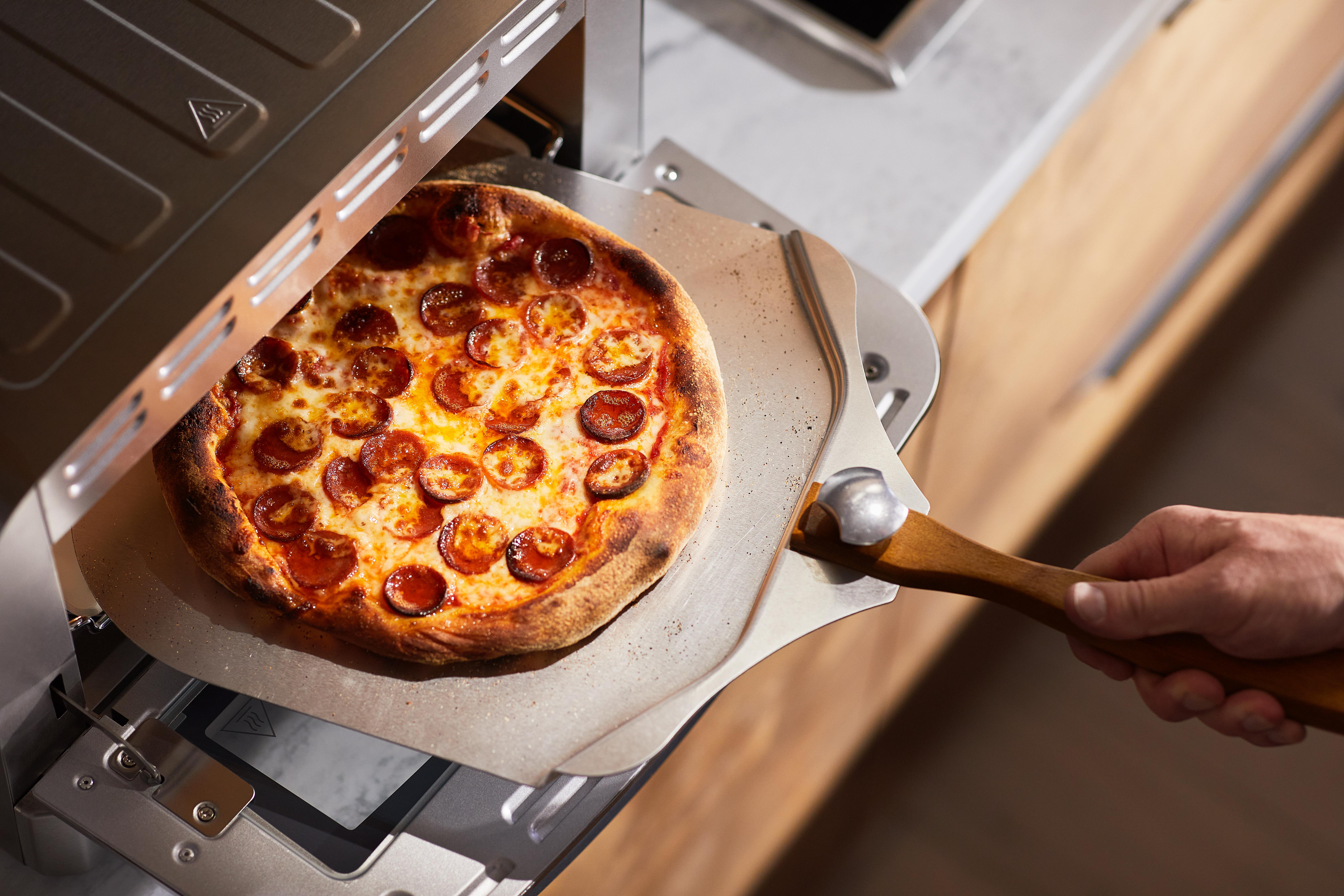 Eine Pizza mit Peperoni wird mit einem Pizzaschieber aus einem Pizzaofen genommen. Die Pizza liegt auf einer Metalloberfläche, gehalten von einem Holzgriff.