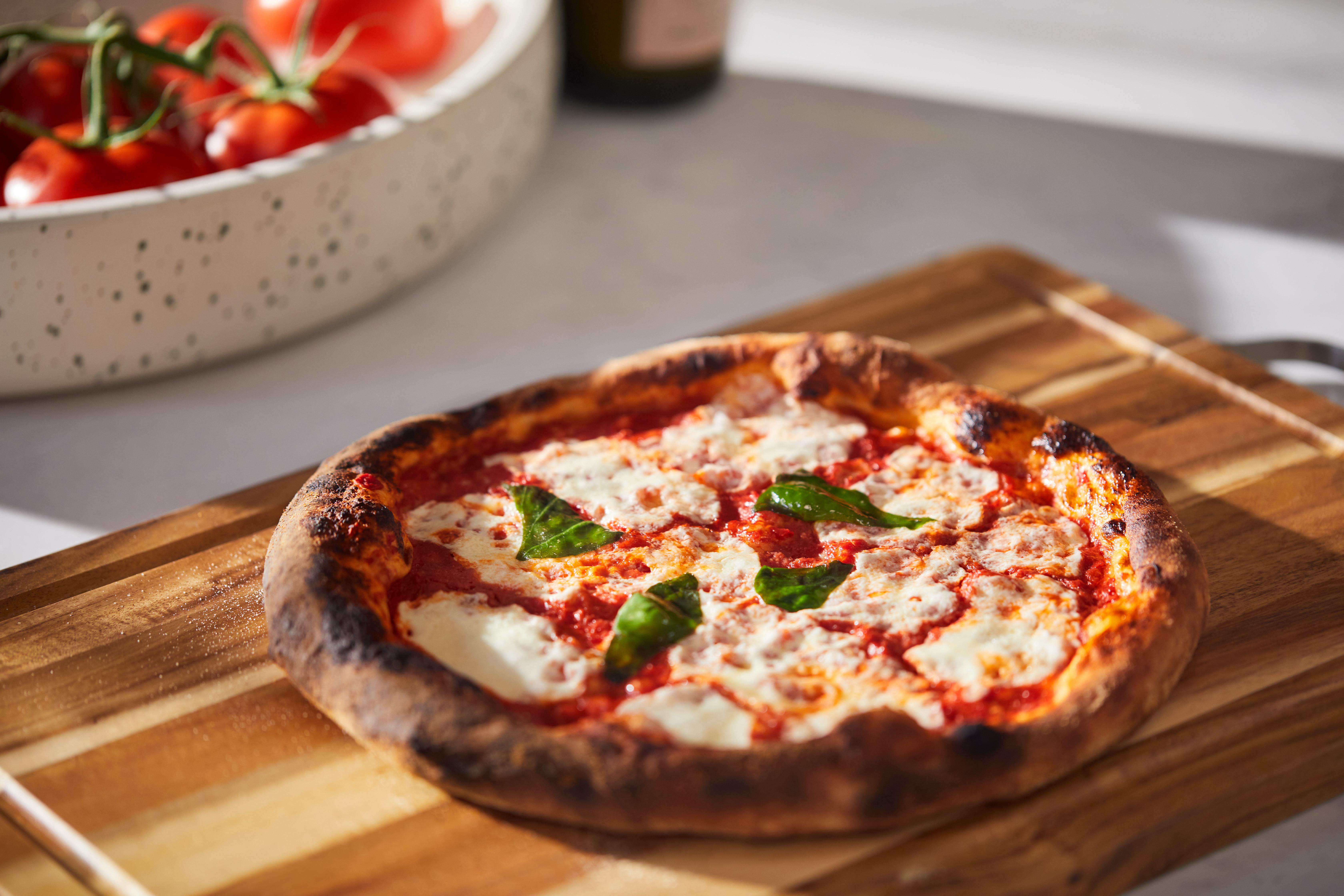 Eine Pizza mit Käse und Basilikum liegt auf einem Holzbrett. Tomaten sind in einer Schüssel im Hintergrund.